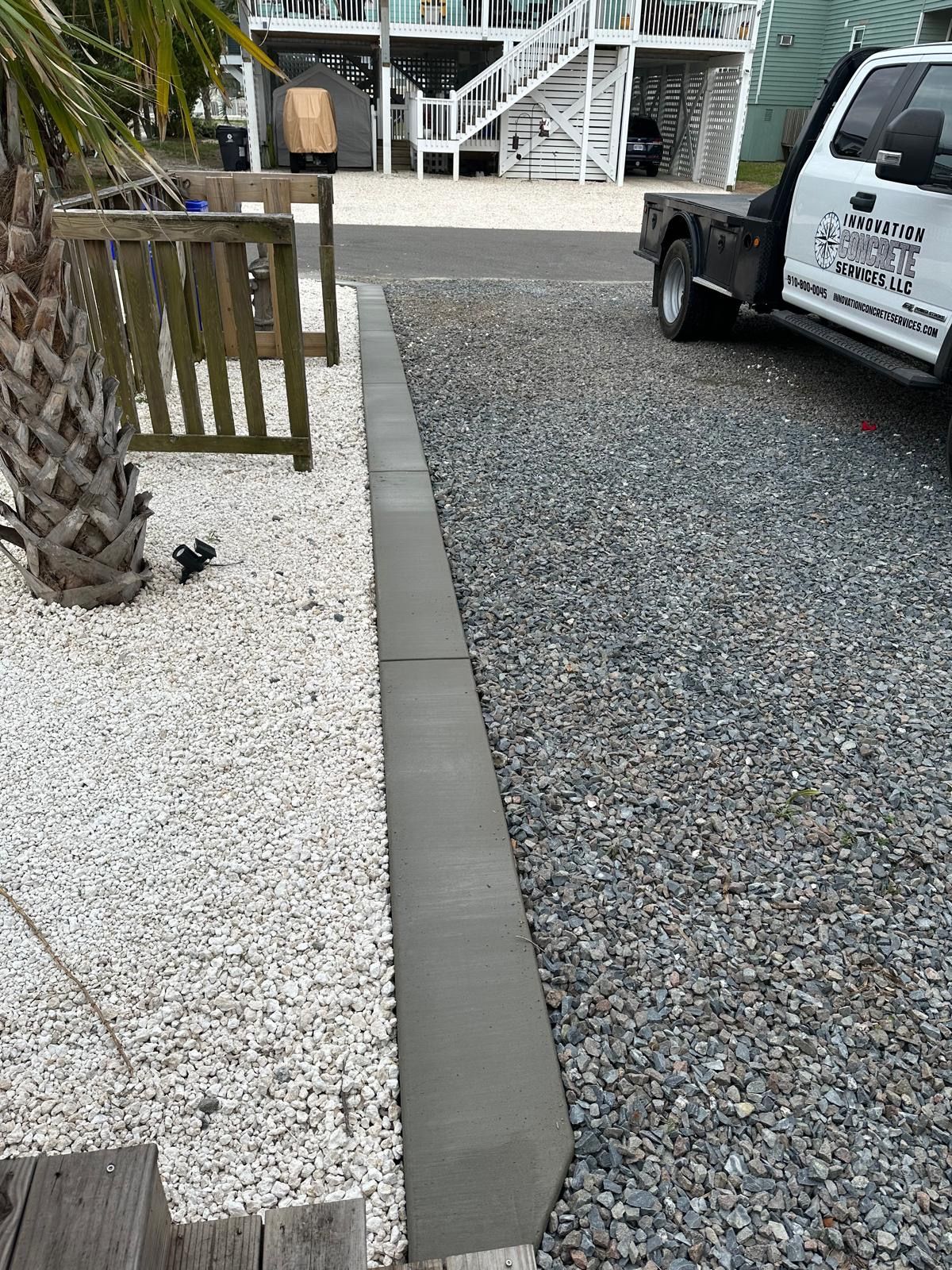 Gravel driveway beside white rock landscaping, with a concrete curb, palm tree, and parked truck near a house.