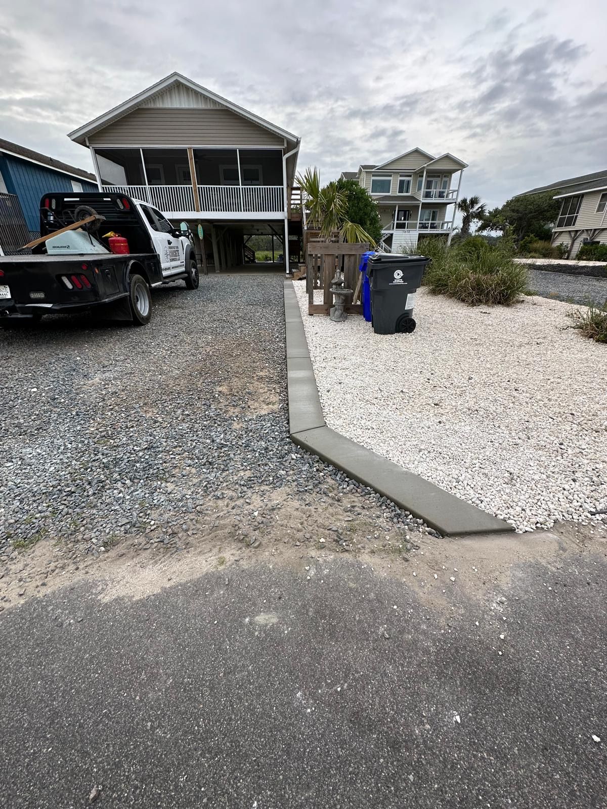 Two houses beside a gravel driveway and white gravel yard, with a black pickup truck and trash bins in front.