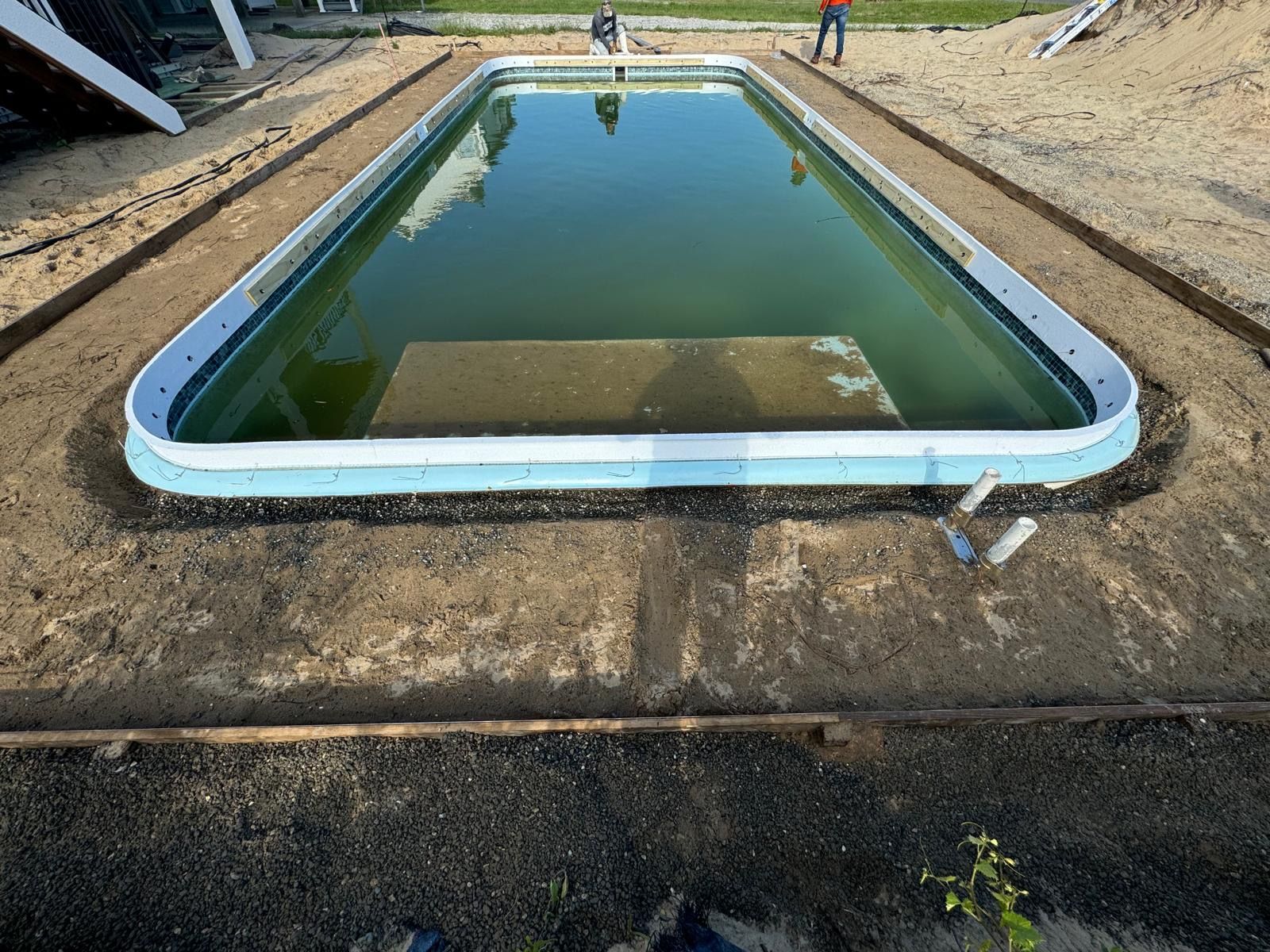 Empty rectangular pool with green water at a construction site, surrounded by dirt and concrete edge