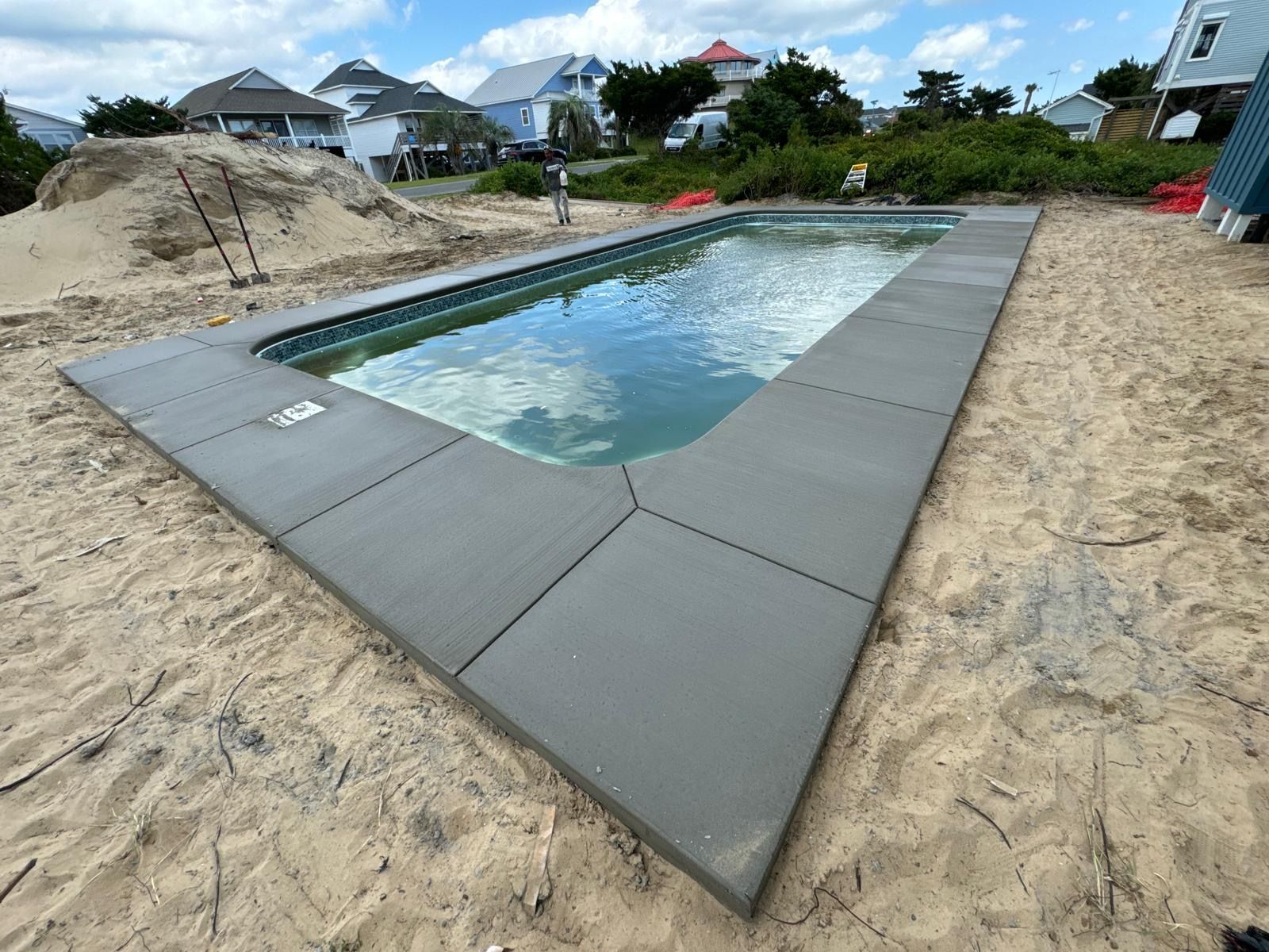 Rectangular outdoor swimming pool with gray deck tiles and sandy surroundings, under a cloudy sky