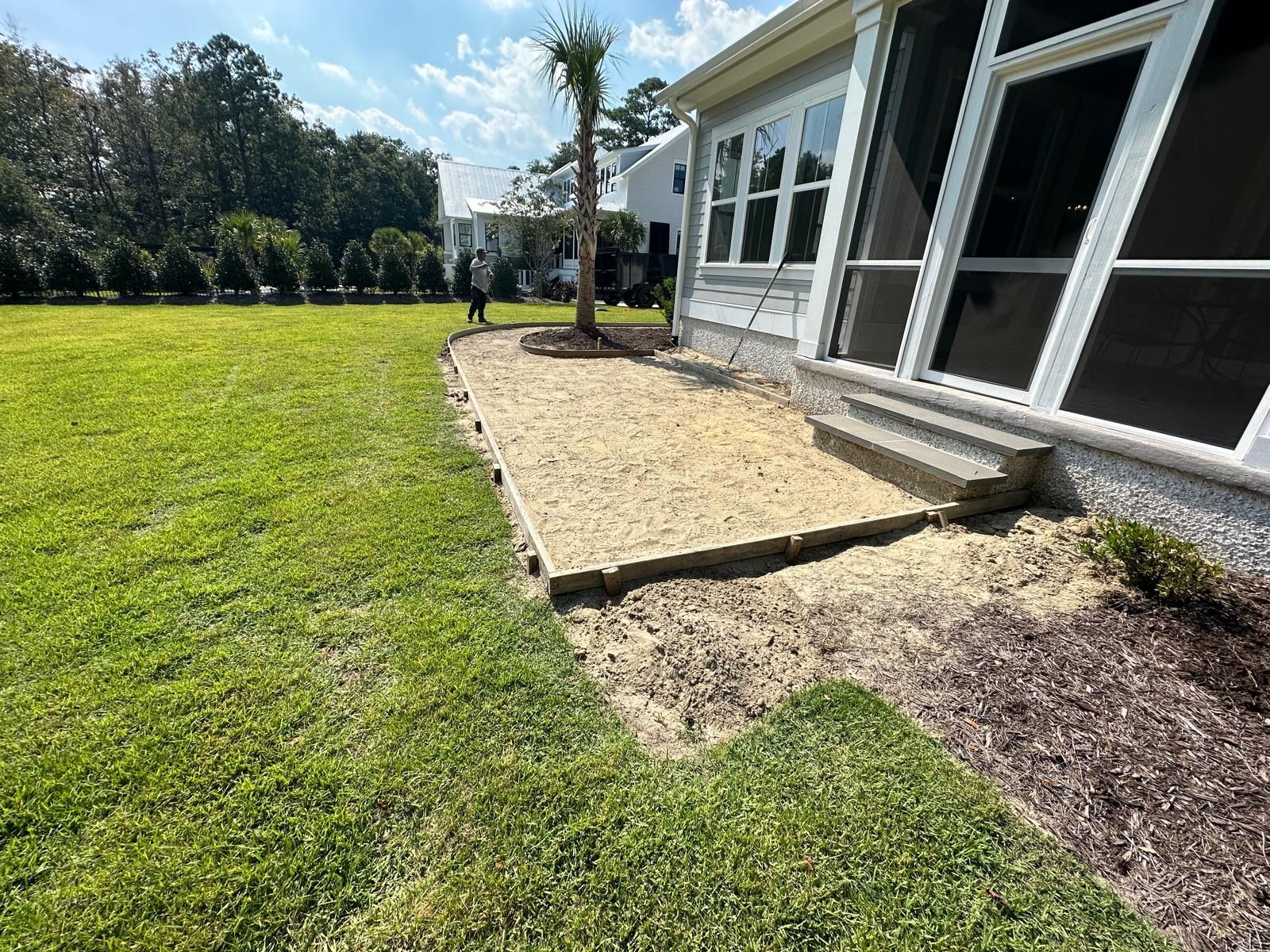 Backyard patio with beige pavers beside a house and a grassy lawn under bright sun