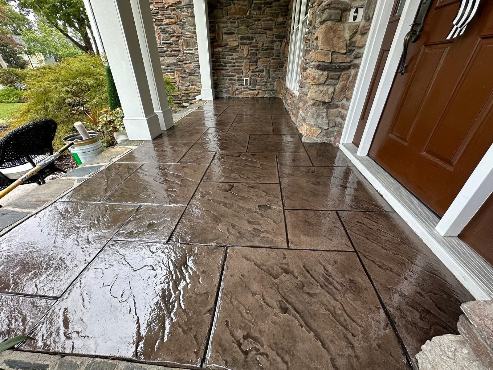 Wet stone porch with brown door, white columns, and green landscaping after rain