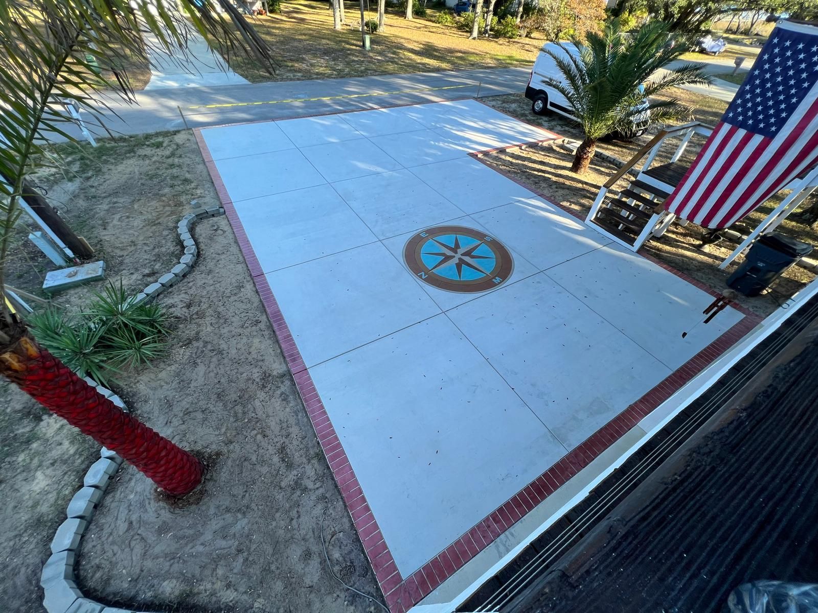 Concrete driveway with a compass design, bordered by red brick, beside a U.S. flag and palm tree.