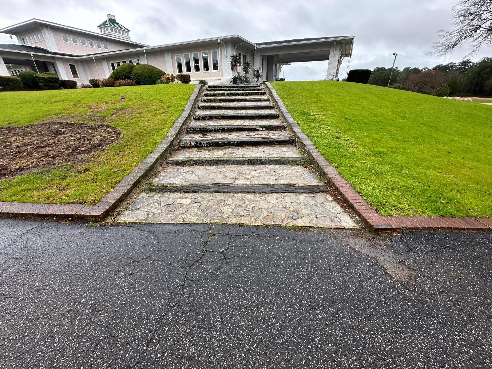 House on a hill with a stone stairway, green lawn, and driveway under cloudy skies