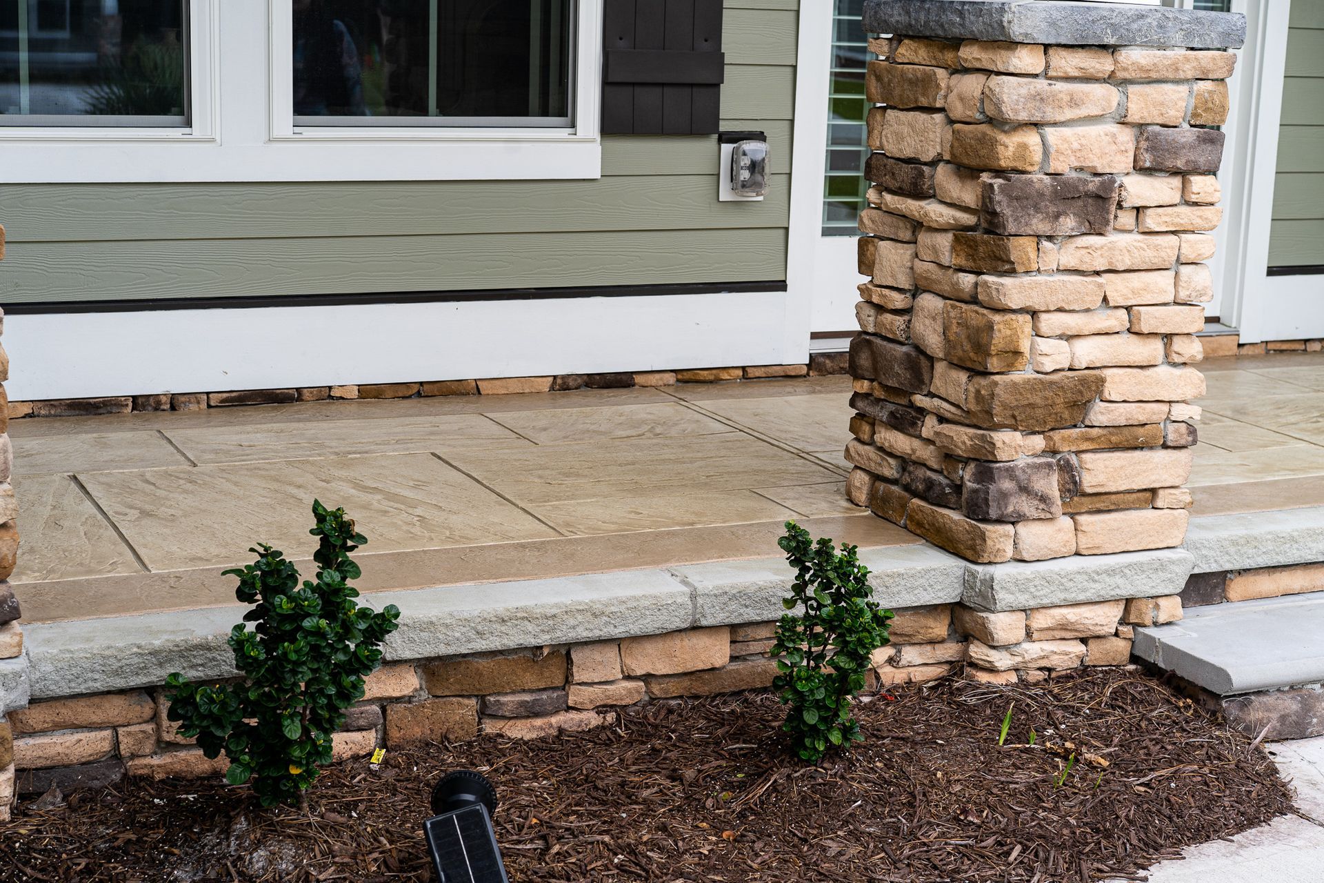 Stone porch and column beside a house exterior with small shrubs and mulch in front