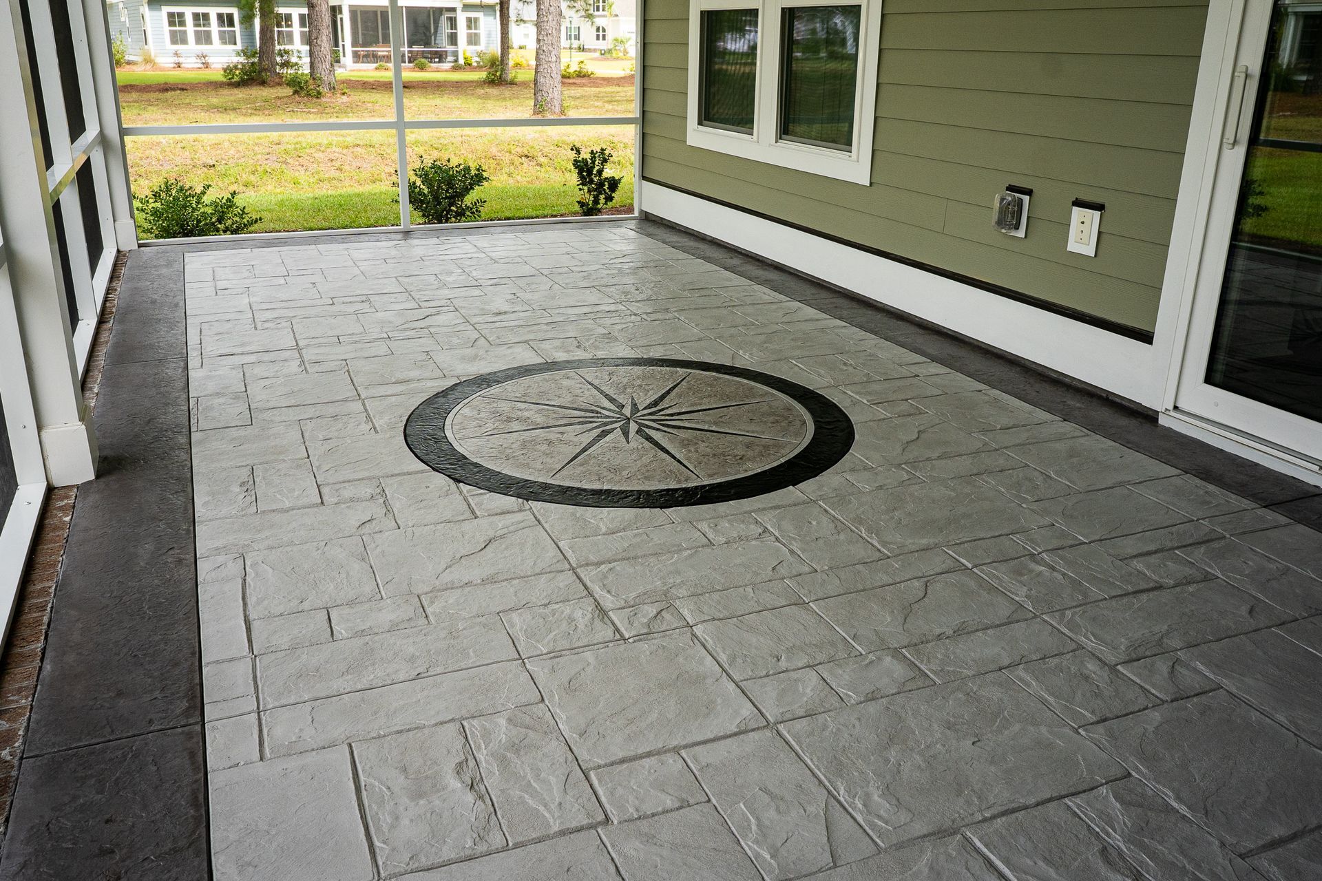 Covered patio with gray stone pavers and a circular inlay, beside a house and yard.