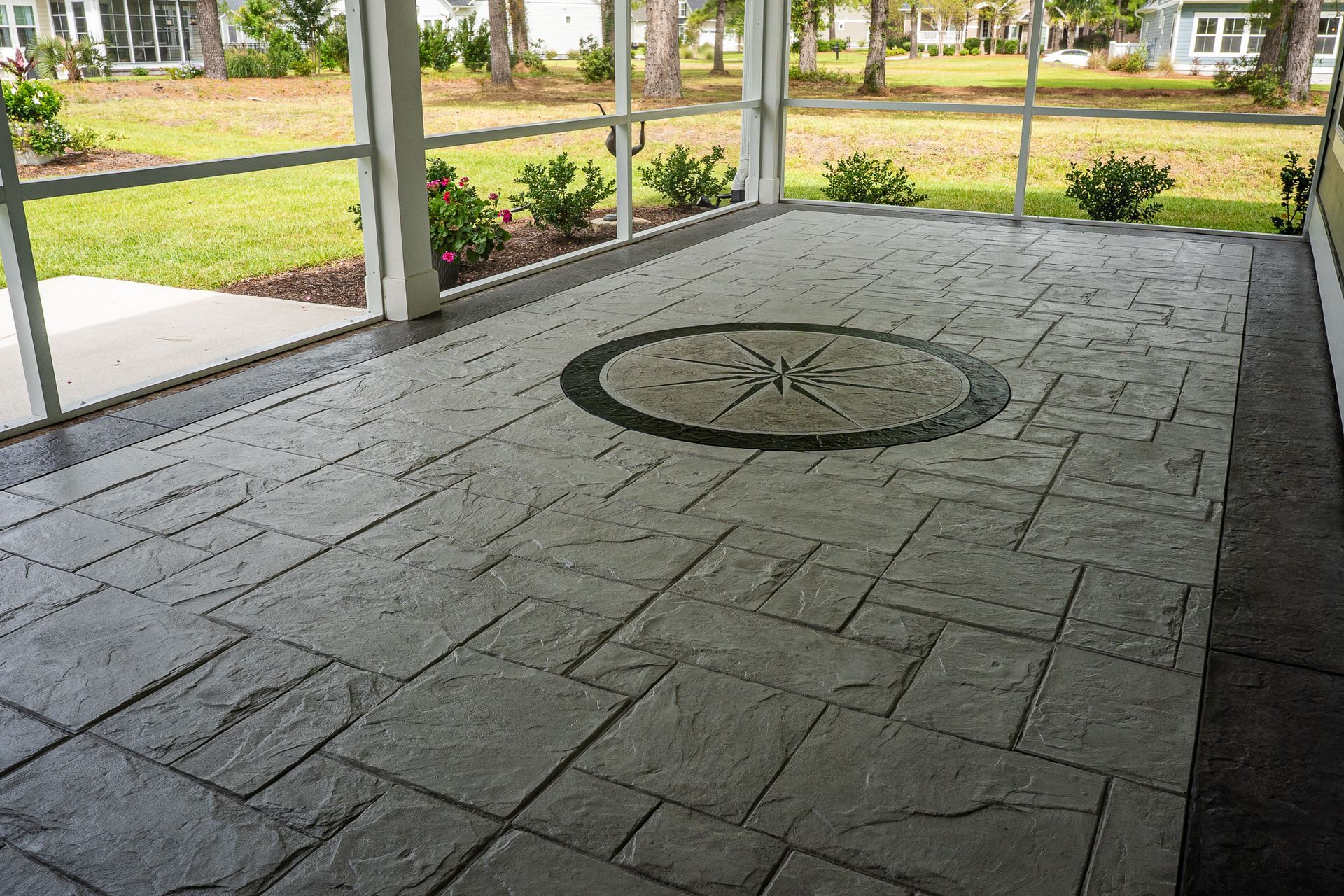 Stone patio with a circular compass design, bordered by screened porch posts and a garden beyond
