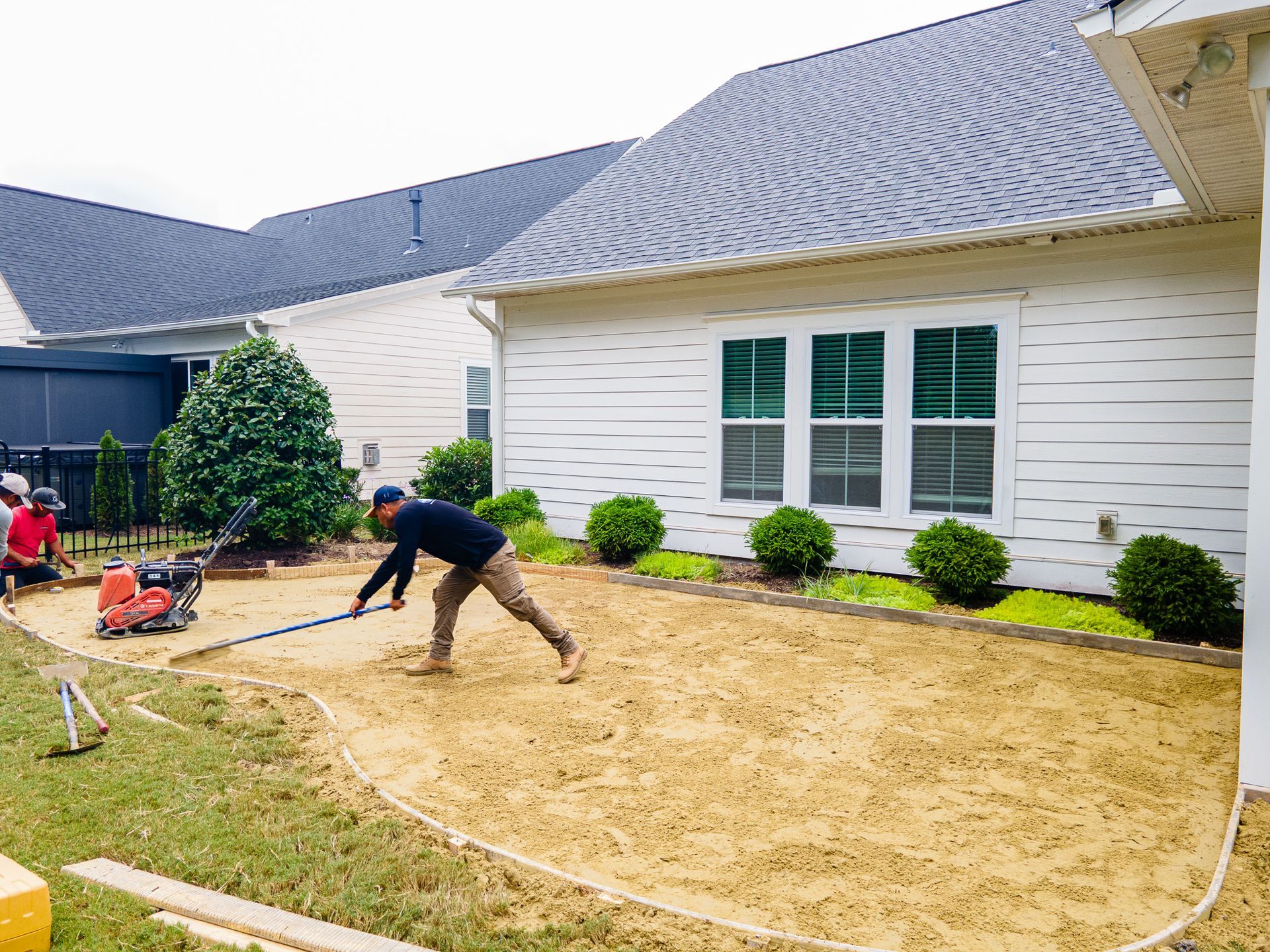 Worker leveling soil in a front yard beside a white house with green-trimmed windows