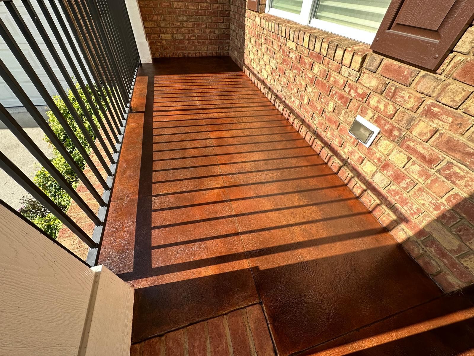 Sunlit apartment balcony with brown wooden floor, black railing, and brick wall
