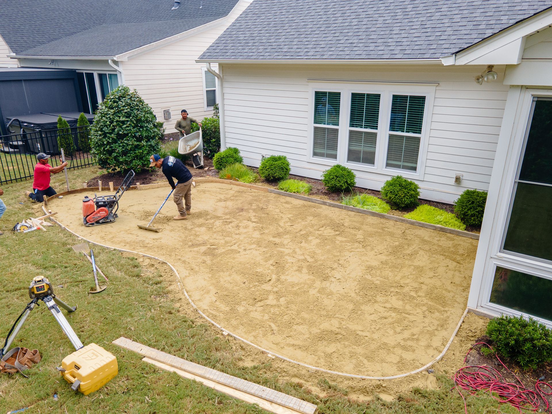 Two workers grading a backyard lawn beside a white house, with tools and hose visible