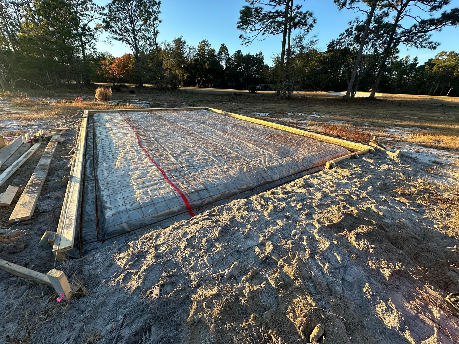 Foundation rebar grid on a sandy construction site at dusk, with lumber and trees nearby