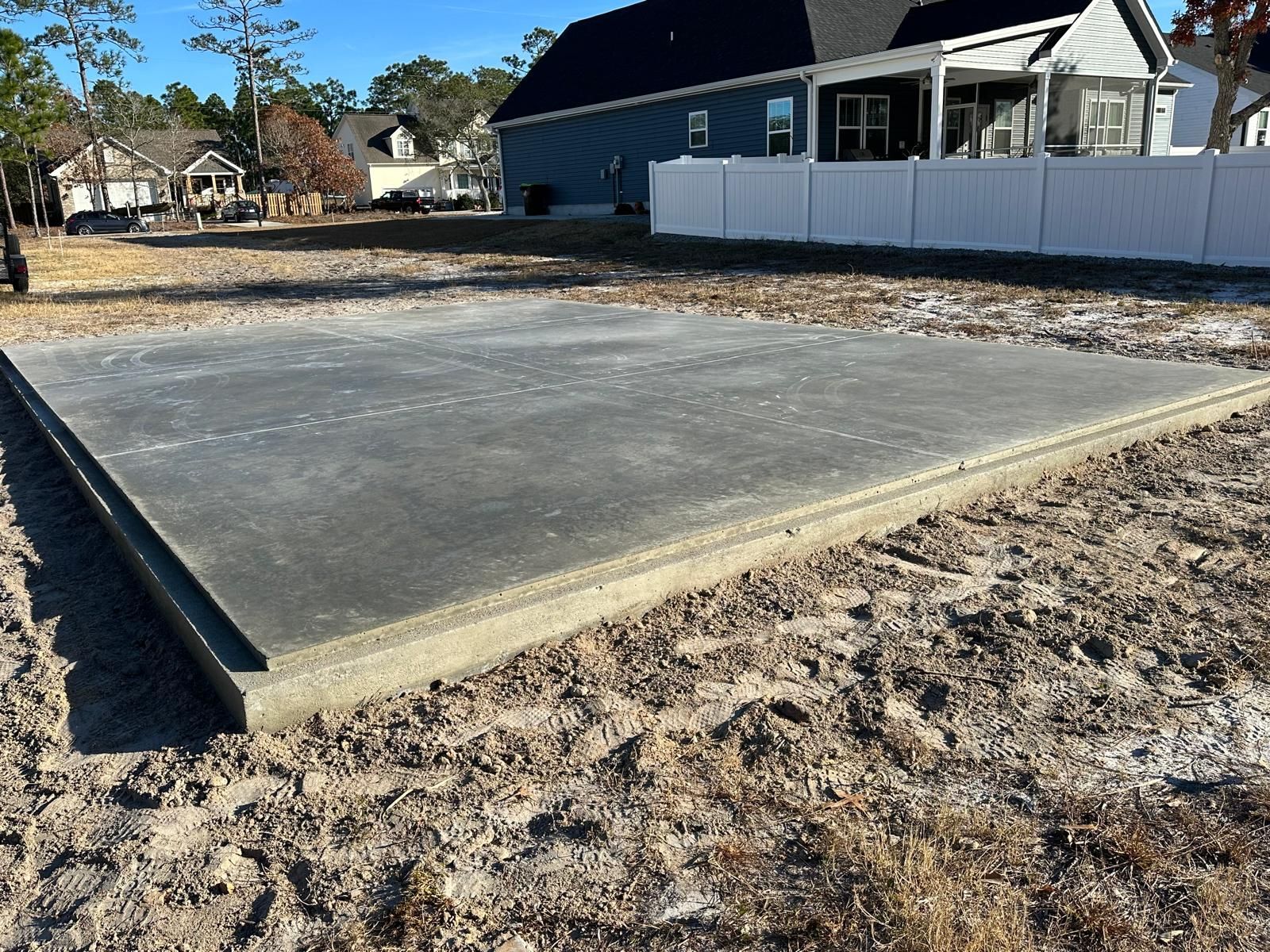 Empty concrete slab in a dirt lot beside a house and white fence on a sunny day