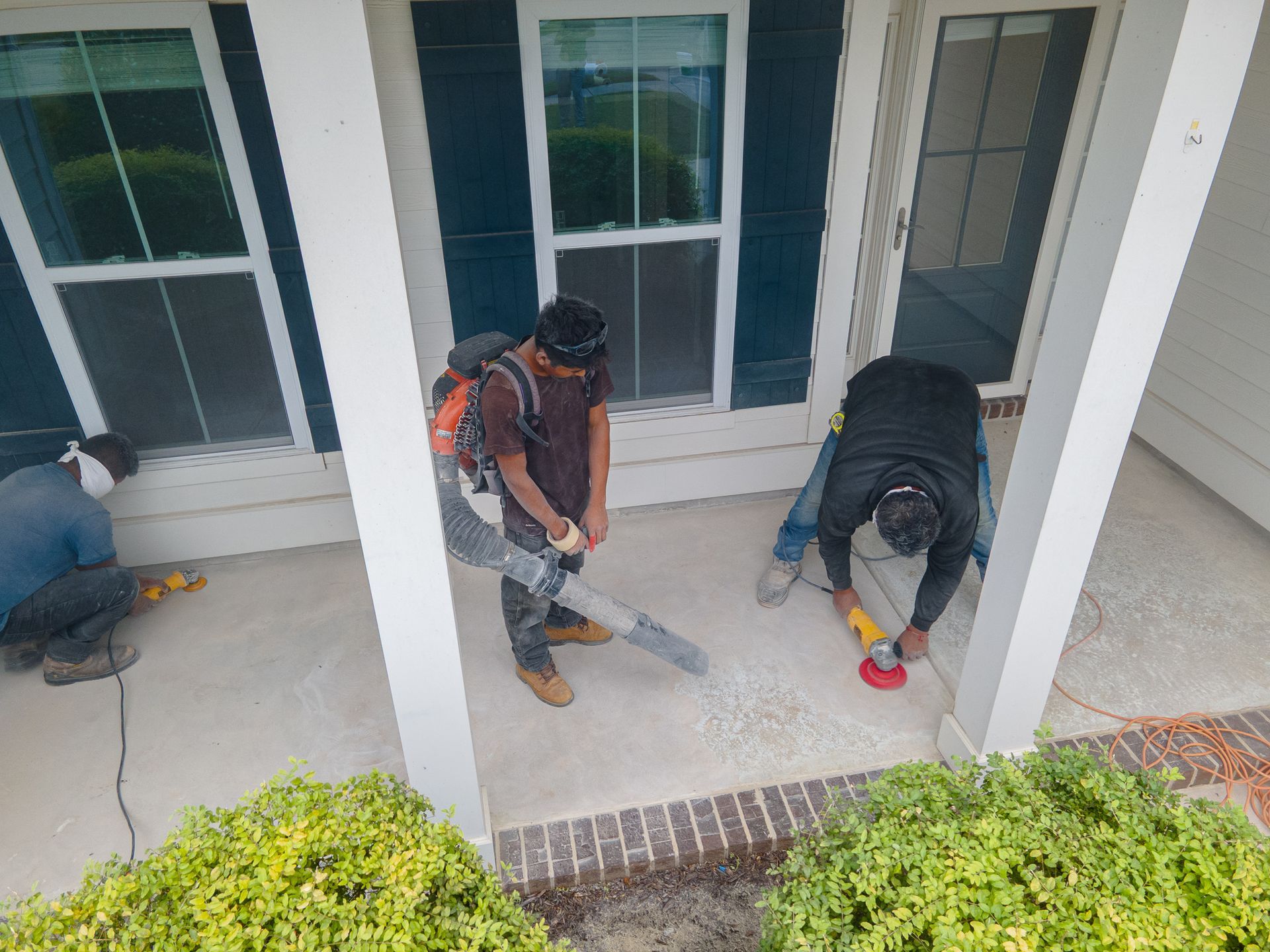 Three workers using power tools to repair concrete on a porch outside a house.