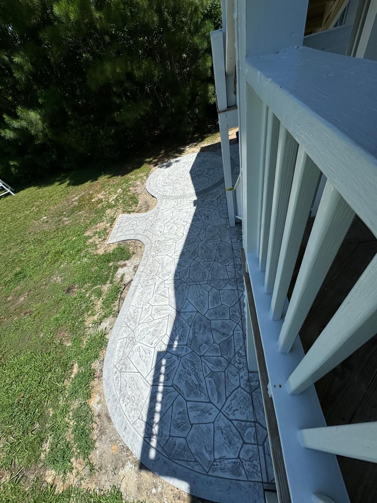 Narrow gravel path beside a white house porch, bordered by grass and trees in sunlight.