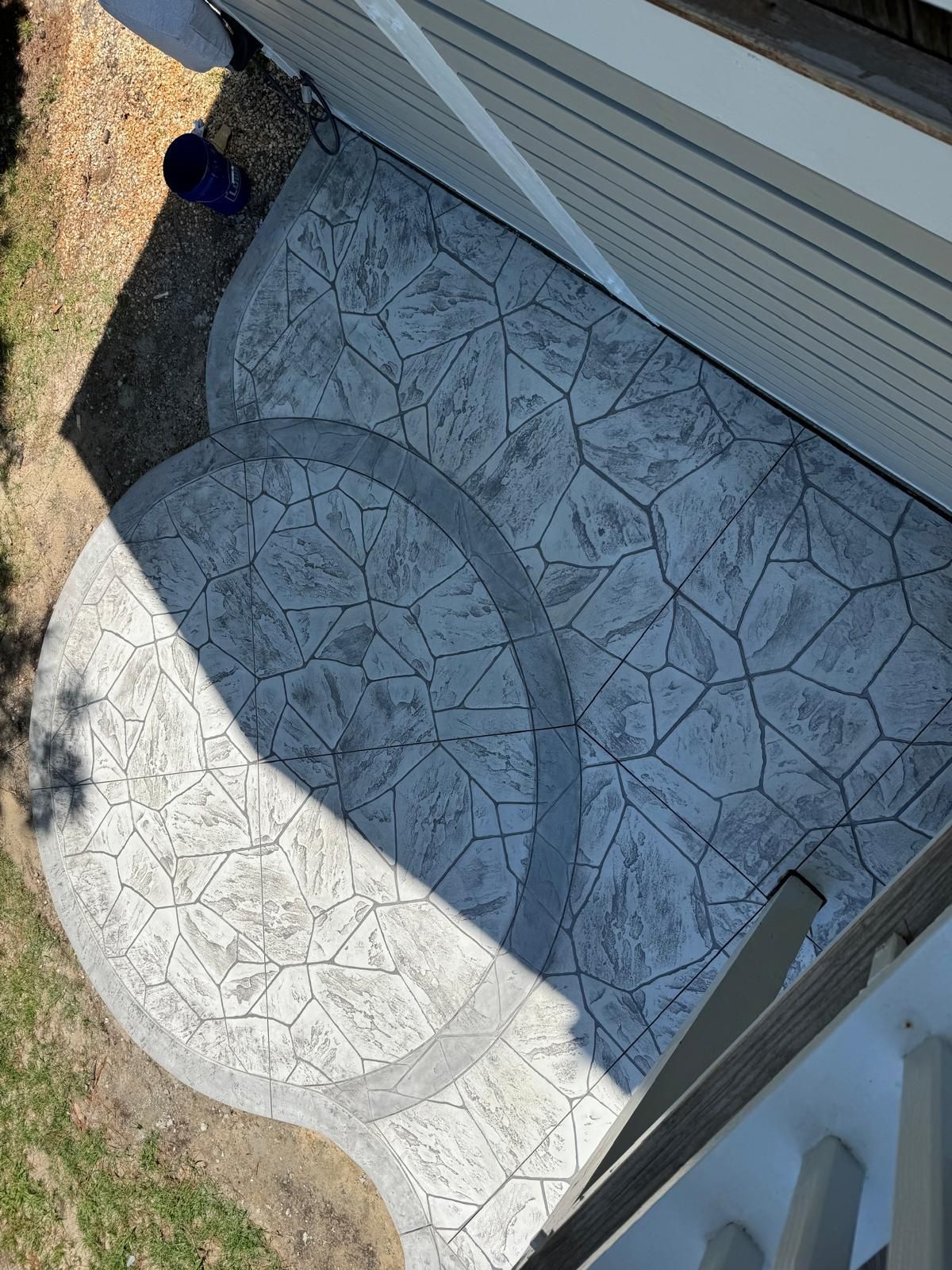 Sunlit stamped concrete patio beside a house, partly shaded by a wall and railing