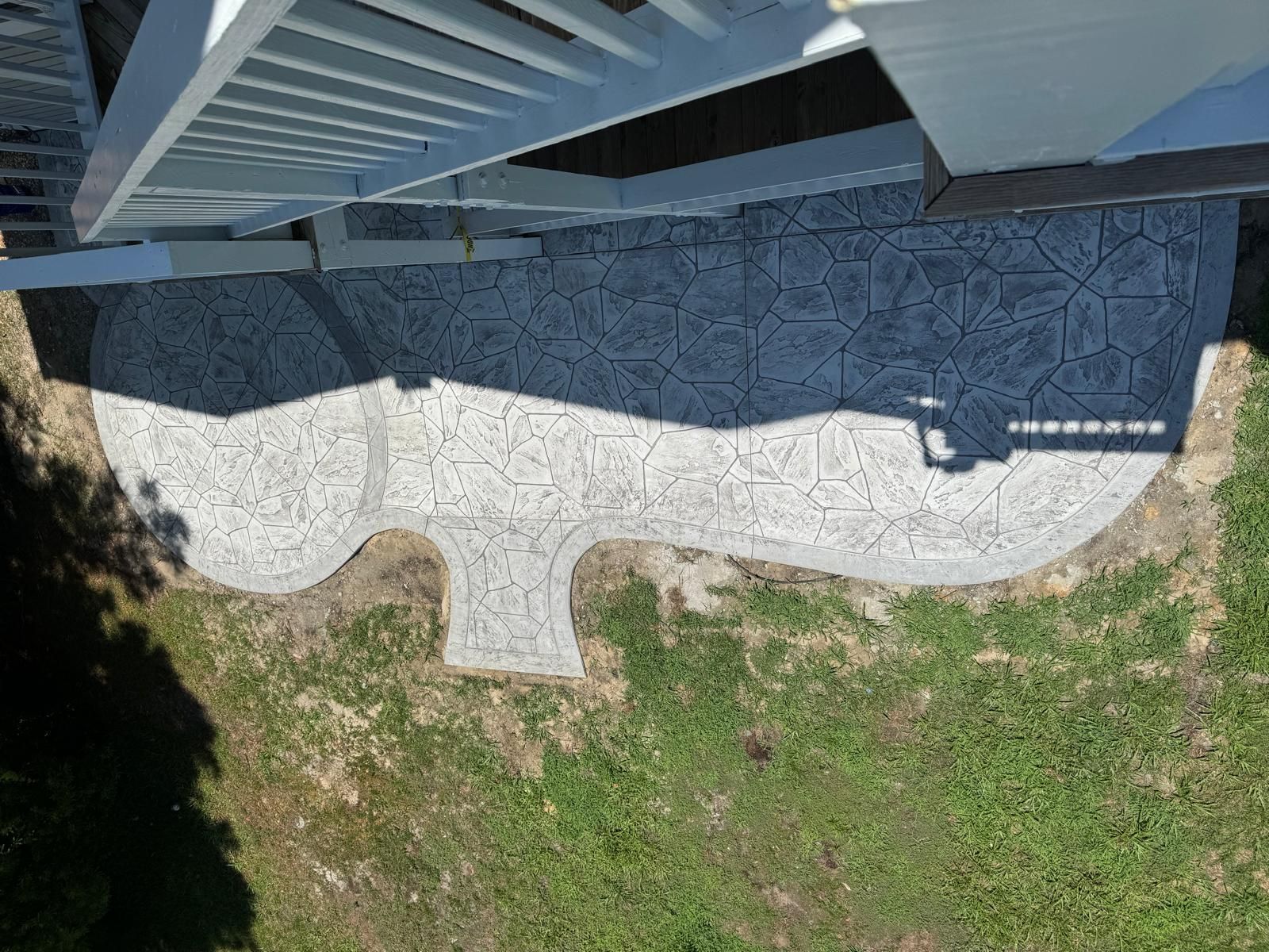 Overhead view of a curved white stone patio beside a house, bordered by grass and shaded by a roof overhang