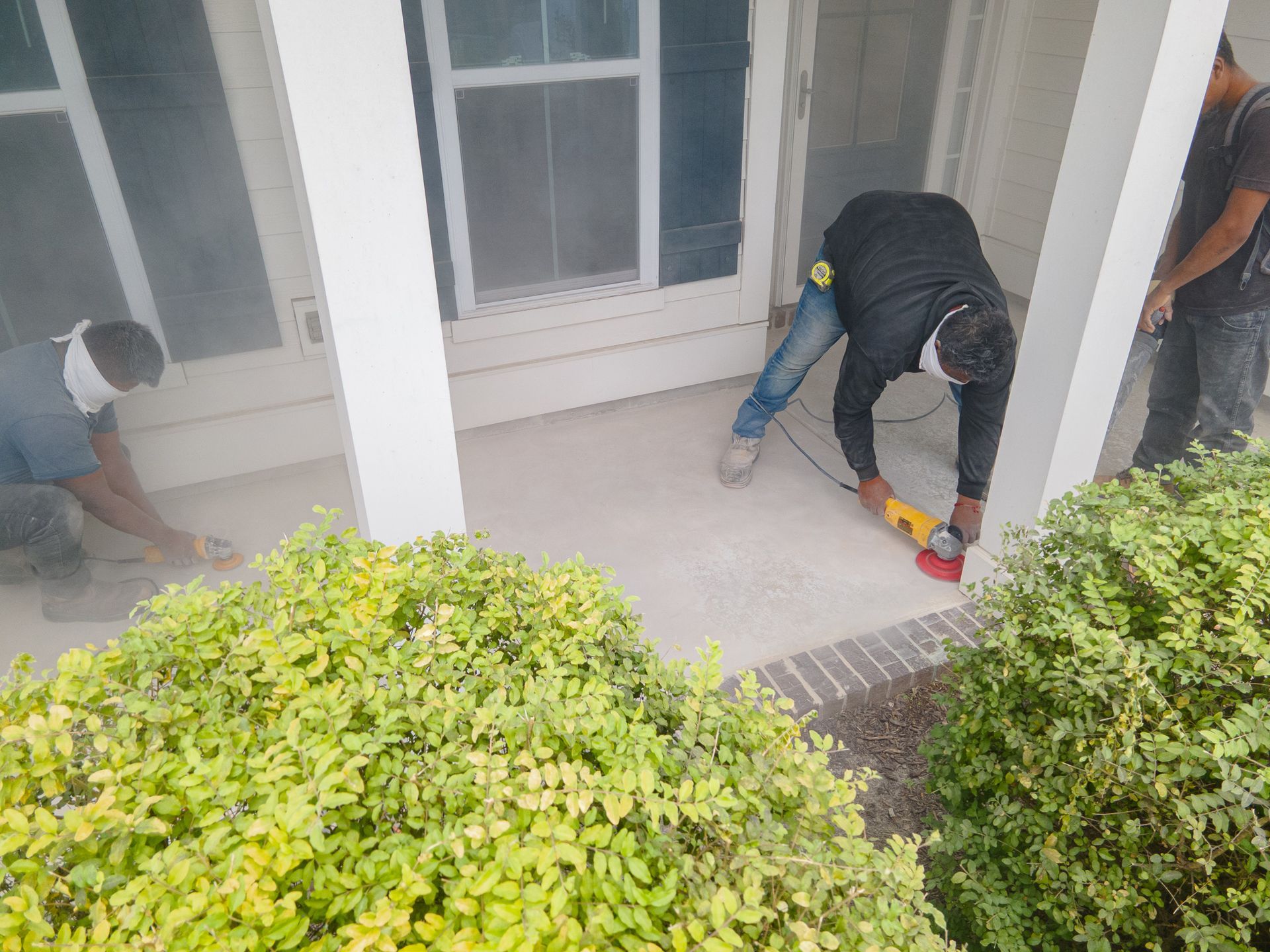 Workers installing flooring on a covered porch beside white French doors and green shrubs