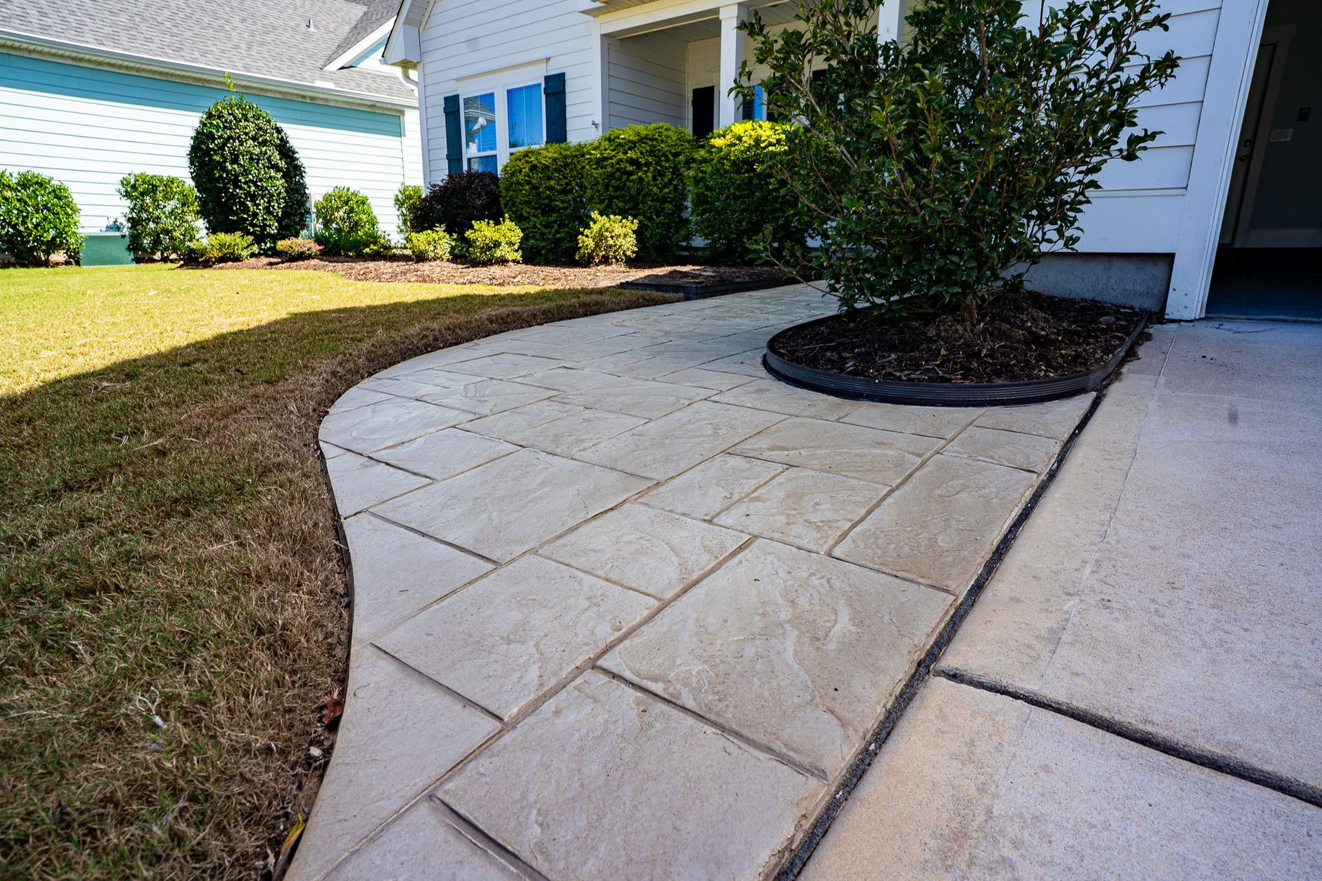 Curved concrete walkway beside a house with trimmed shrubs and a grassy lawn