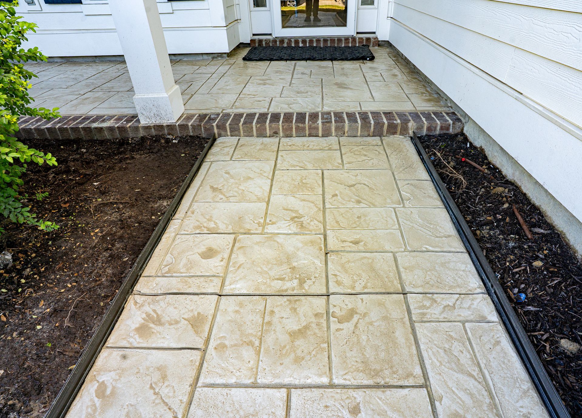 Tiled front walkway leading to a house porch, bordered by dark mulch and white siding