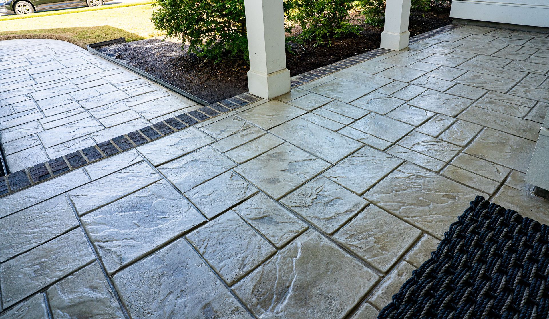 Covered stone patio with wet textured pavers, a white column, and a mat by the entrance.