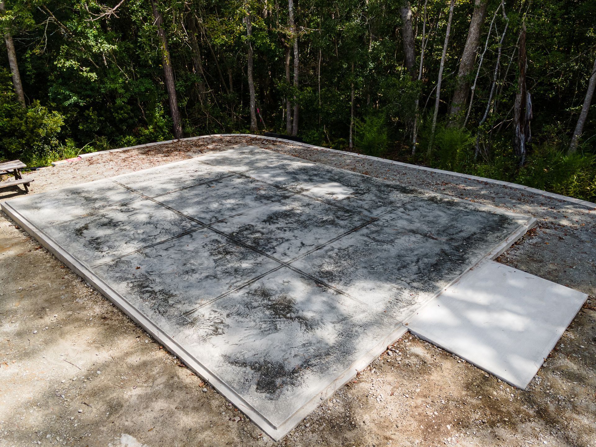 Concrete slab foundation under construction in a wooded area, with a white panel on one corner.