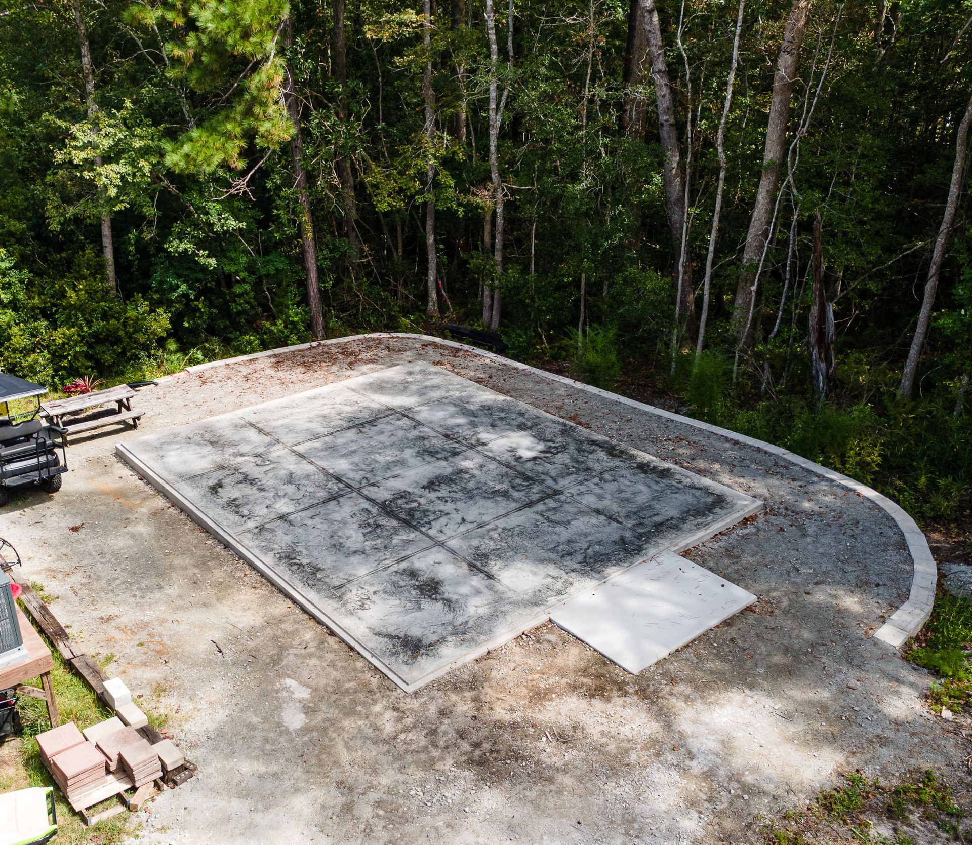 Concrete slab foundation under construction beside a wooded area, with dirt and stacked materials nearby