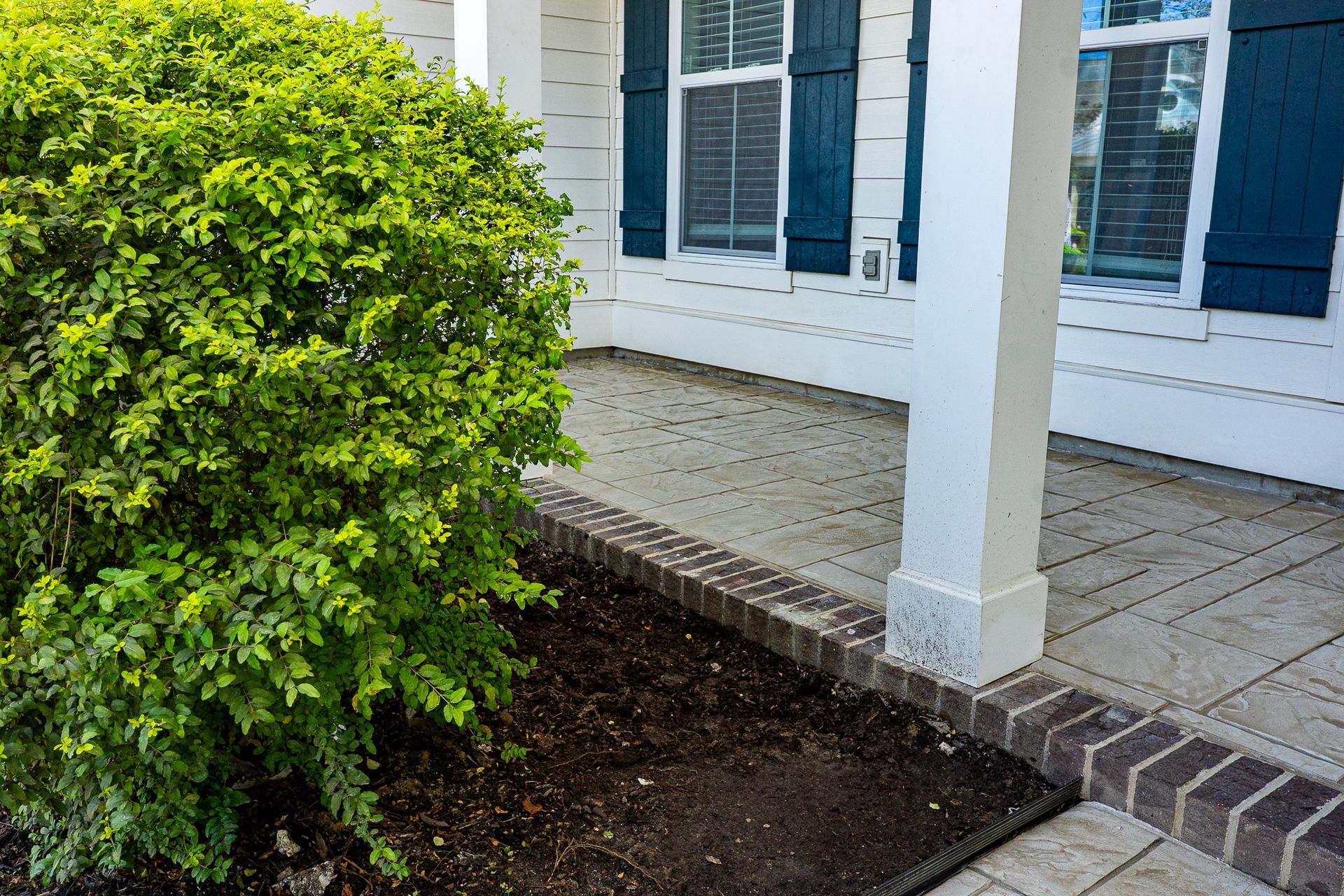 Front porch with white siding, blue shutters, brick steps, and a green shrub in the foreground