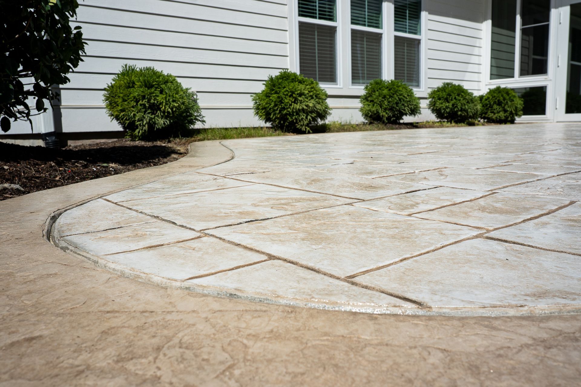 Curved stamped concrete patio beside a white house with shrubs and windows