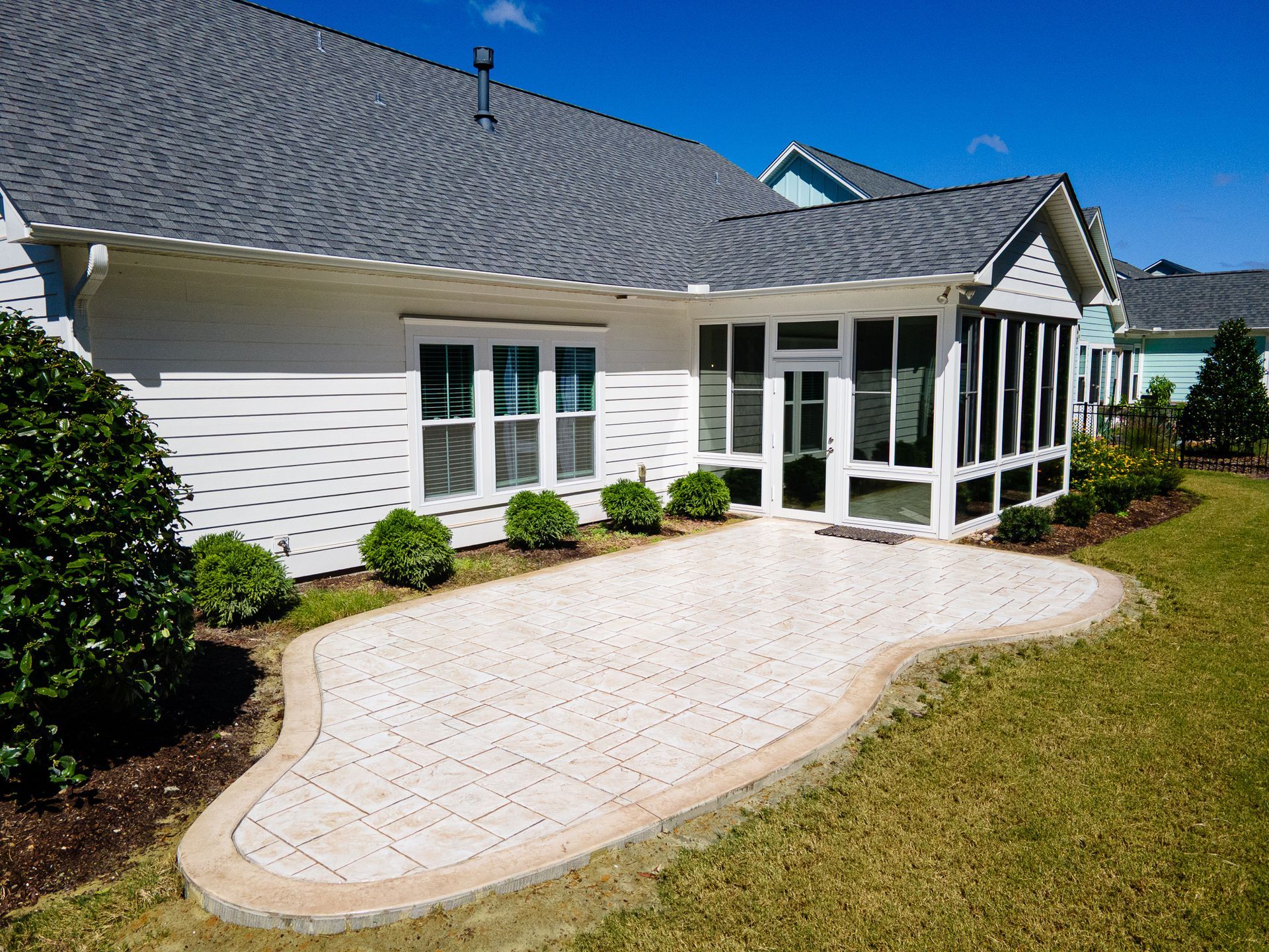 White house with gray roof, sunroom, and curved brick patio beside a lawn and shrubs