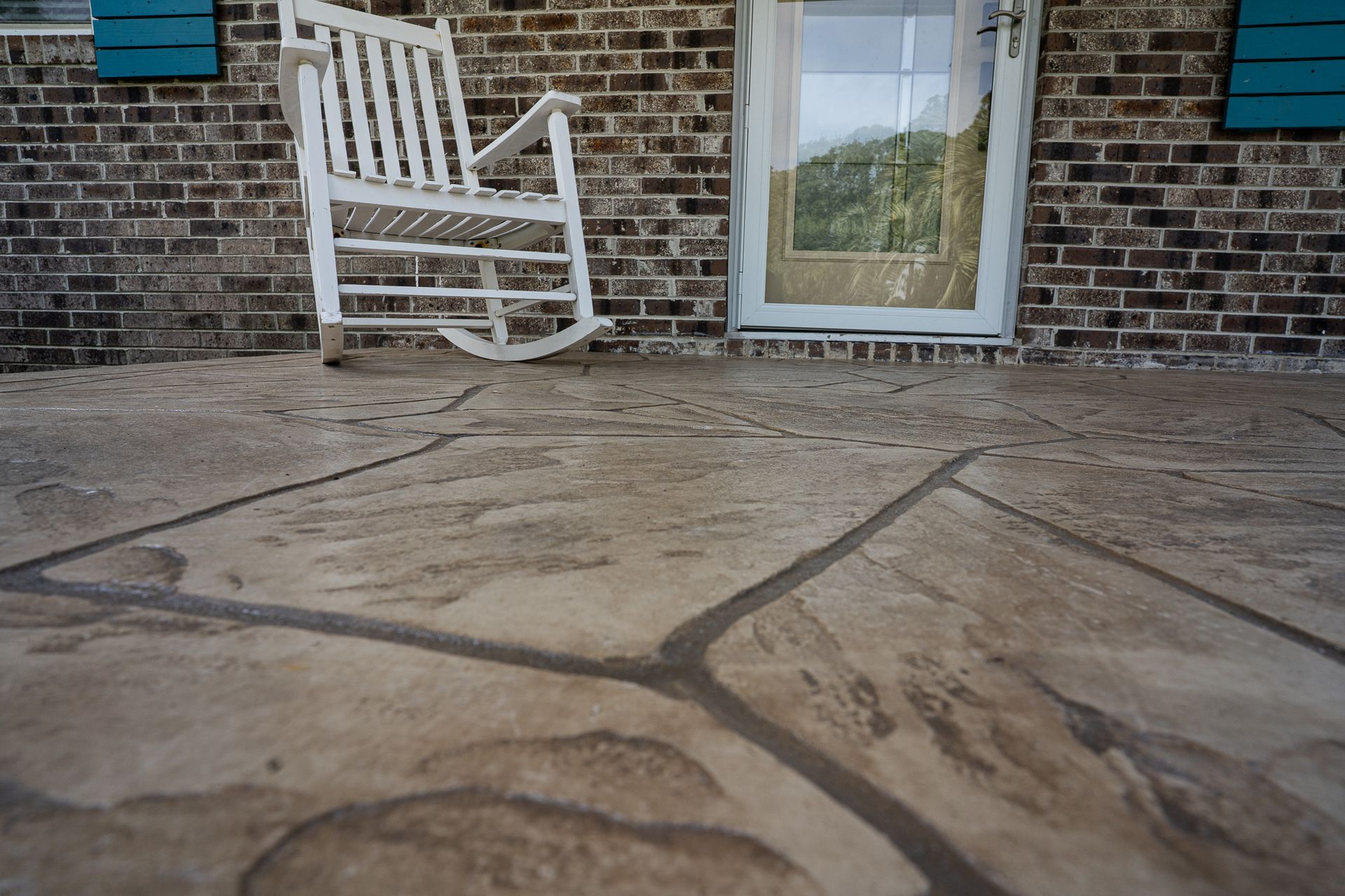 White rocking chair on a brick porch beside a glass door, with stone pavers in the foreground
