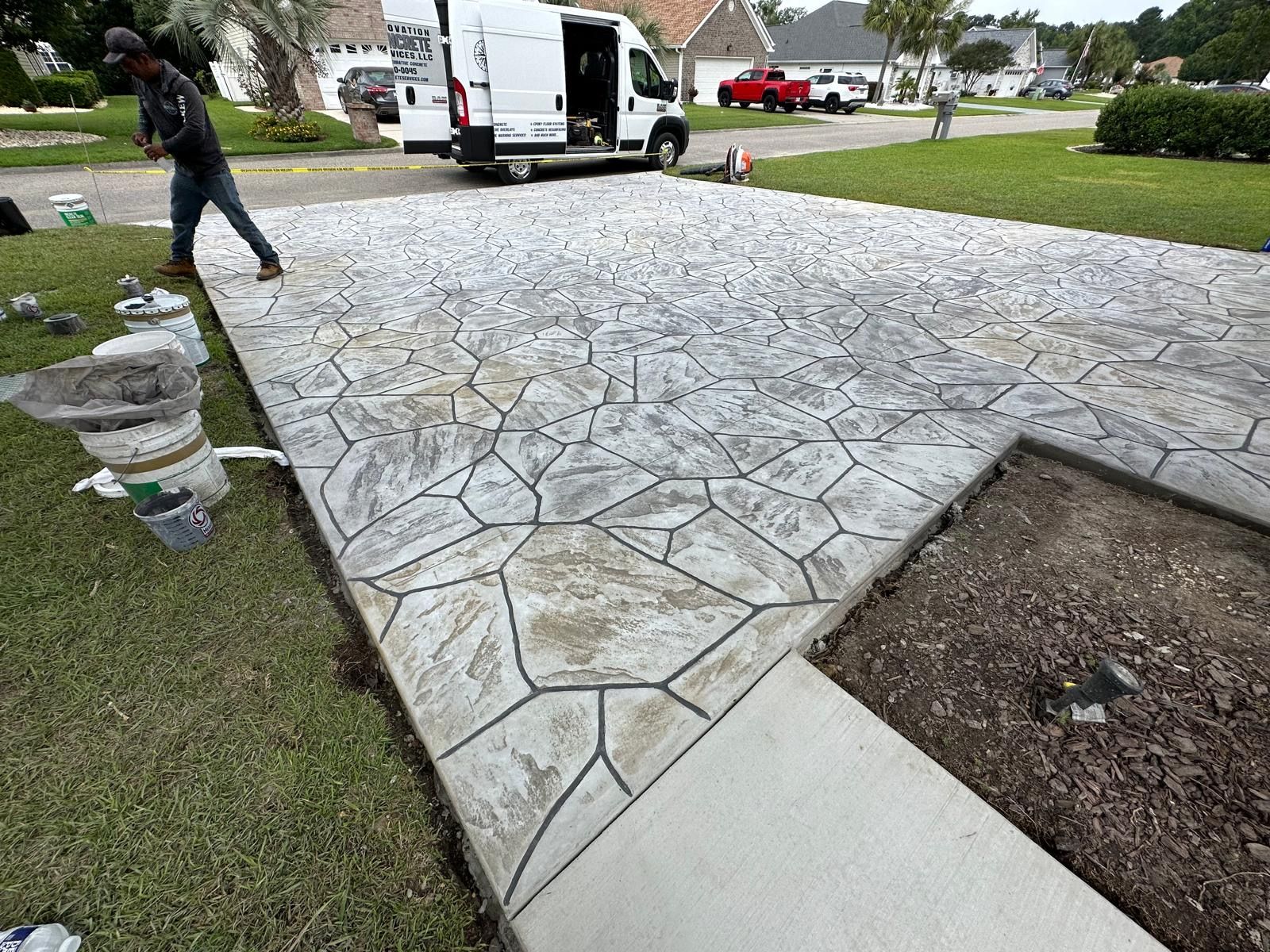Stained concrete driveway being cleaned with a pressure washer beside a white van and lawn.