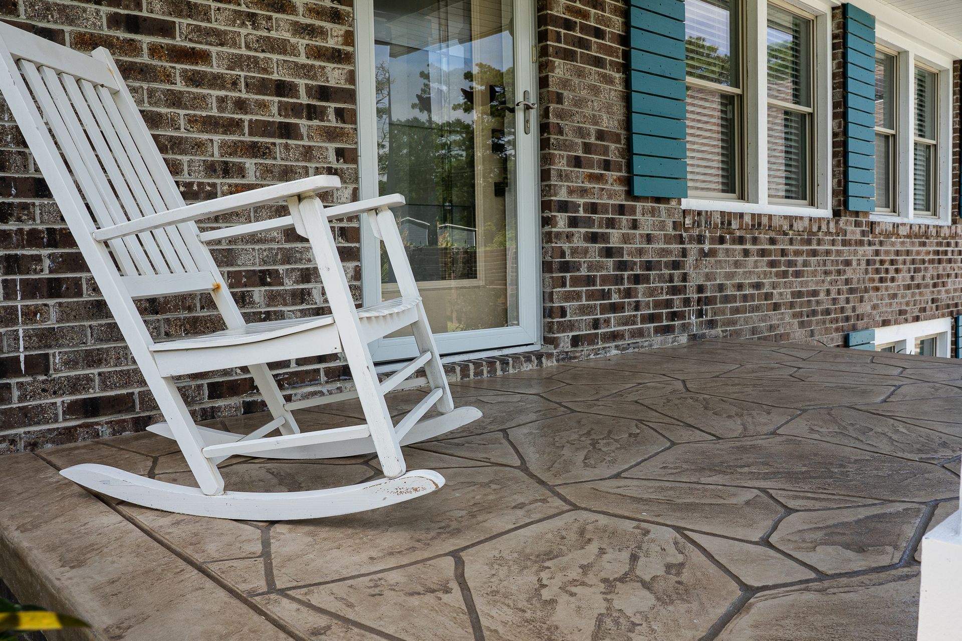 White rocking chair on a stone patio beside a brick house with teal shutters.