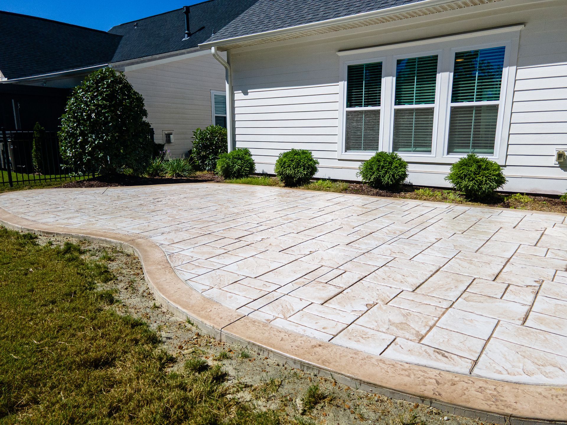 Curved stone patio beside a white house with windows, shrubs, and a grassy lawn