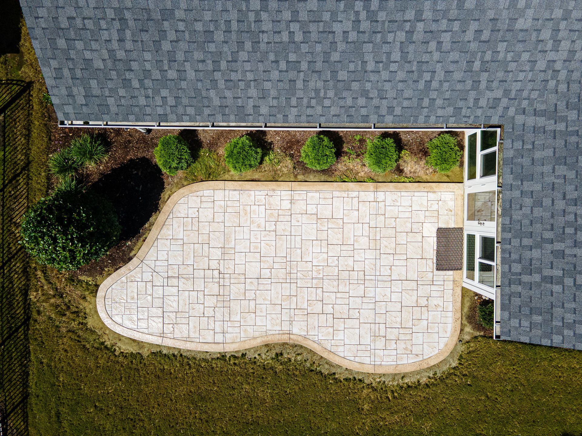 Aerial view of a paved patio beside a house with gray shingles and surrounding grass and shrubs