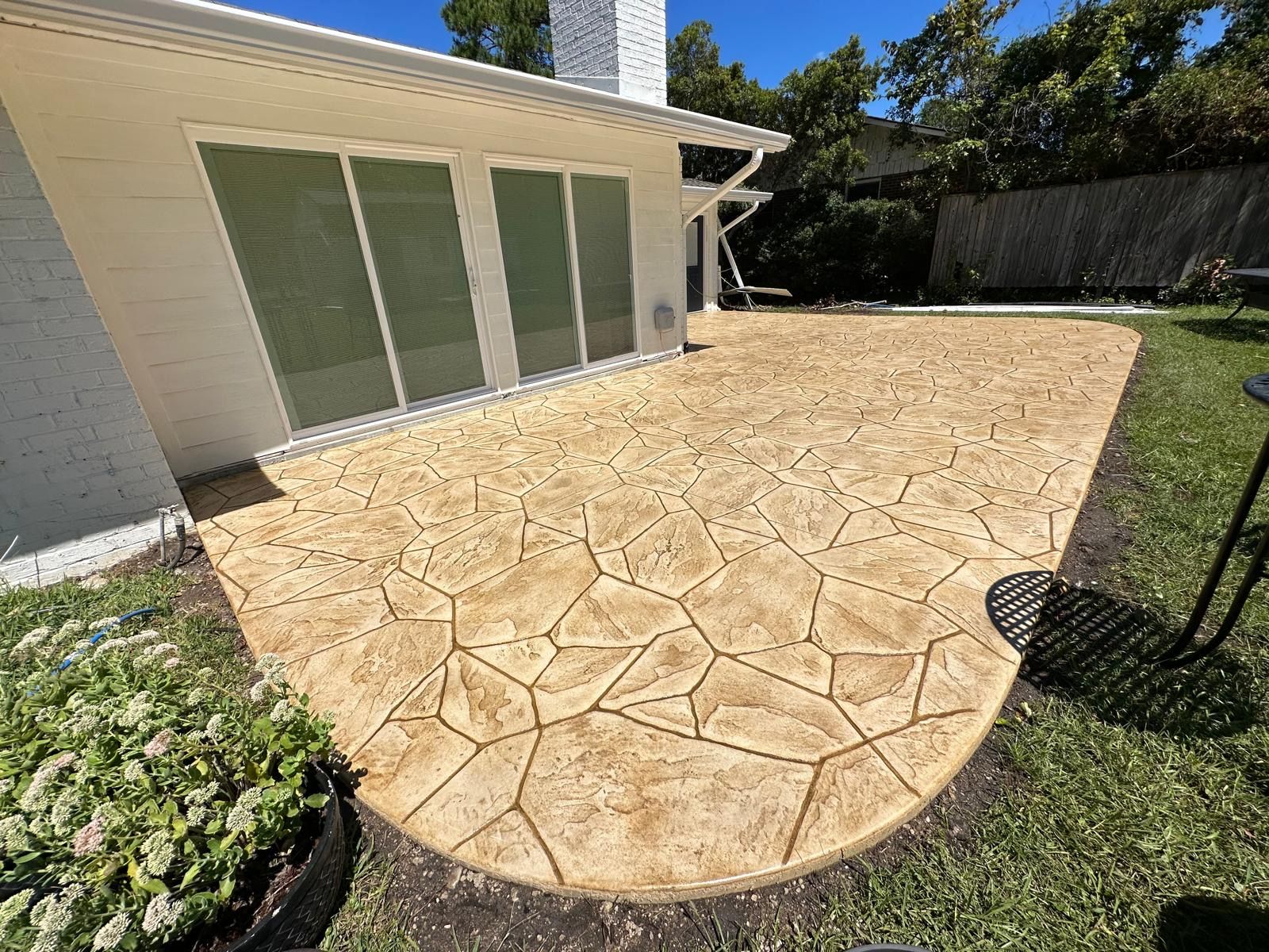 Patio with tan stamped concrete beside a house, bordered by grass and a black fence.