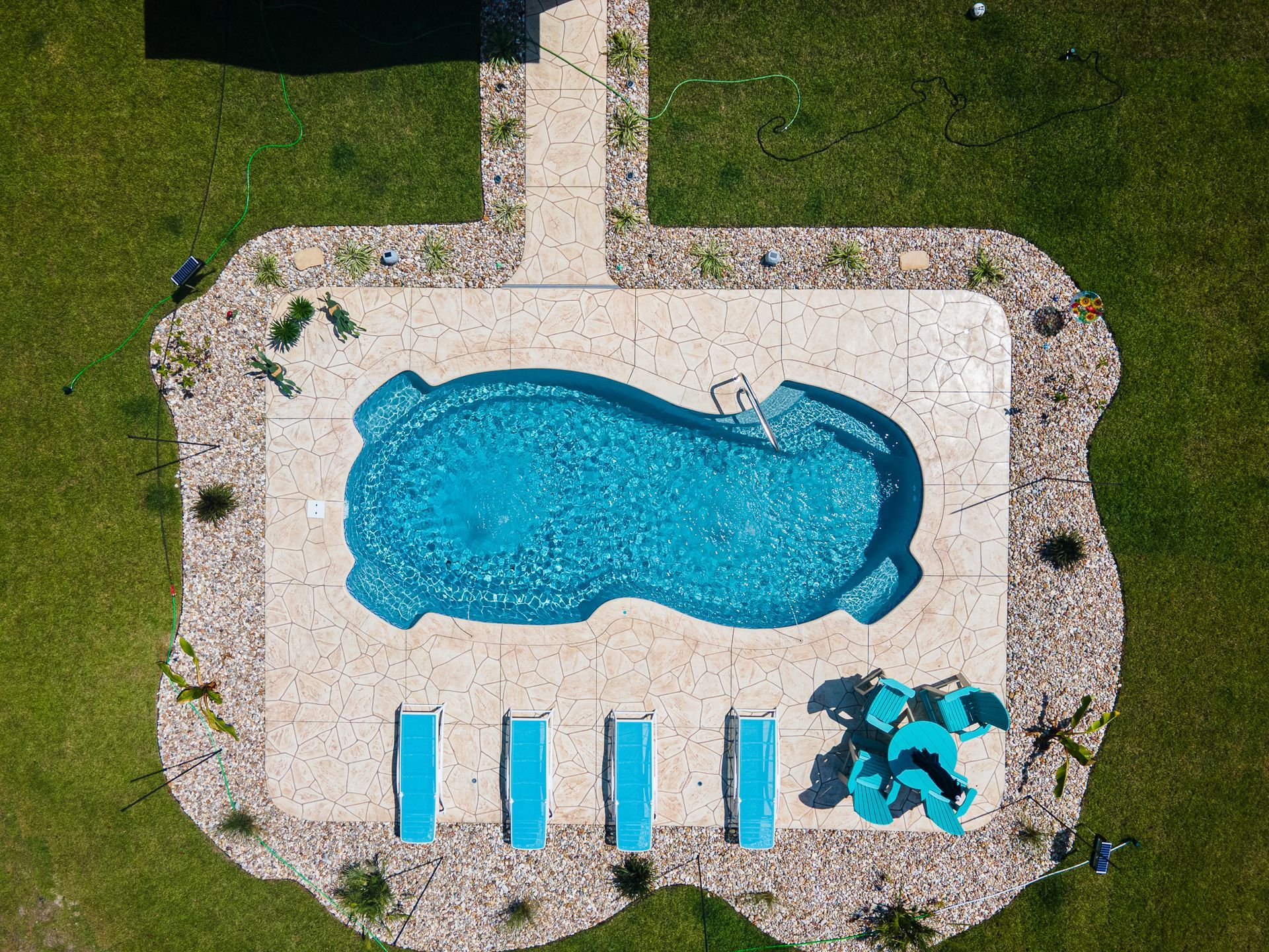 Aerial view of a backyard pool with blue water, white patio, lounge chairs, and green lawn.