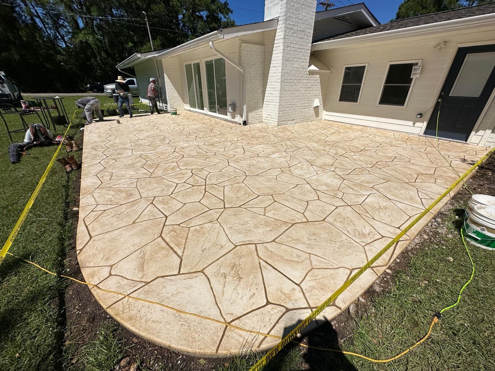 Stone patio under construction beside a house, with workers and hoses around the yard.