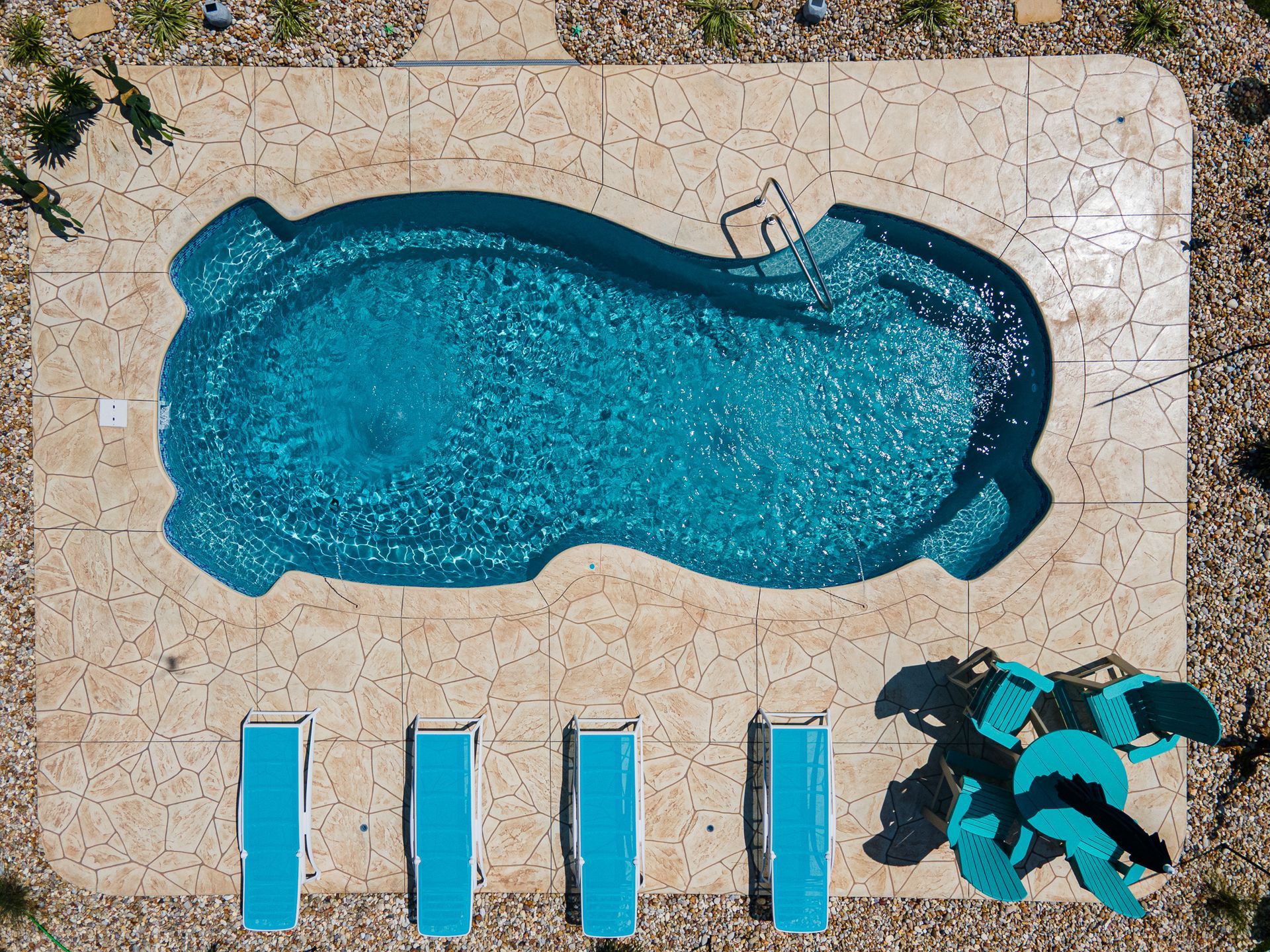 Overhead view of a backyard pool with turquoise water, four lounge chairs, and teal umbrellas on a patio.