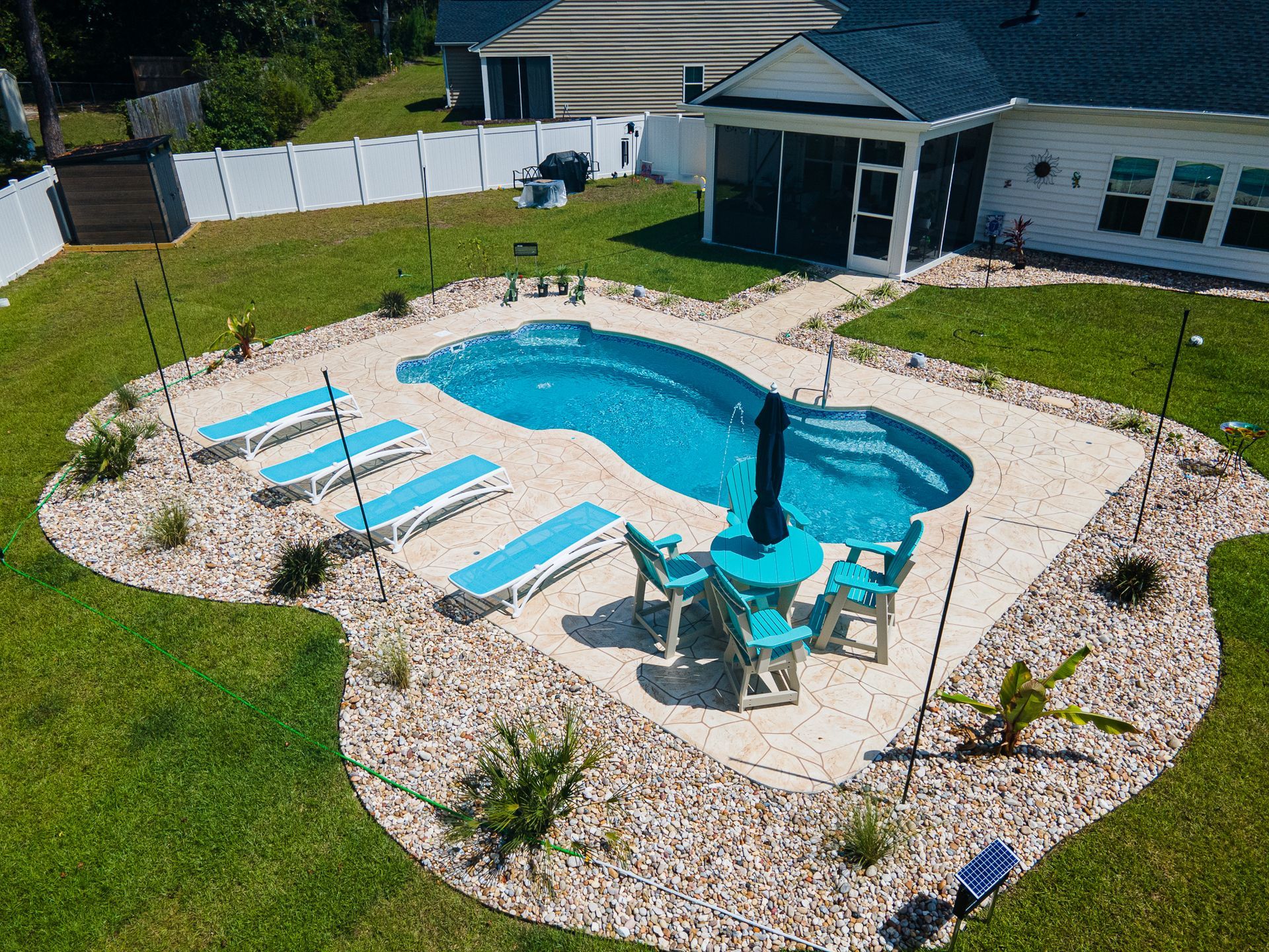 Backyard swimming pool with lounge chairs and blue umbrellas, surrounded by gravel and grass.