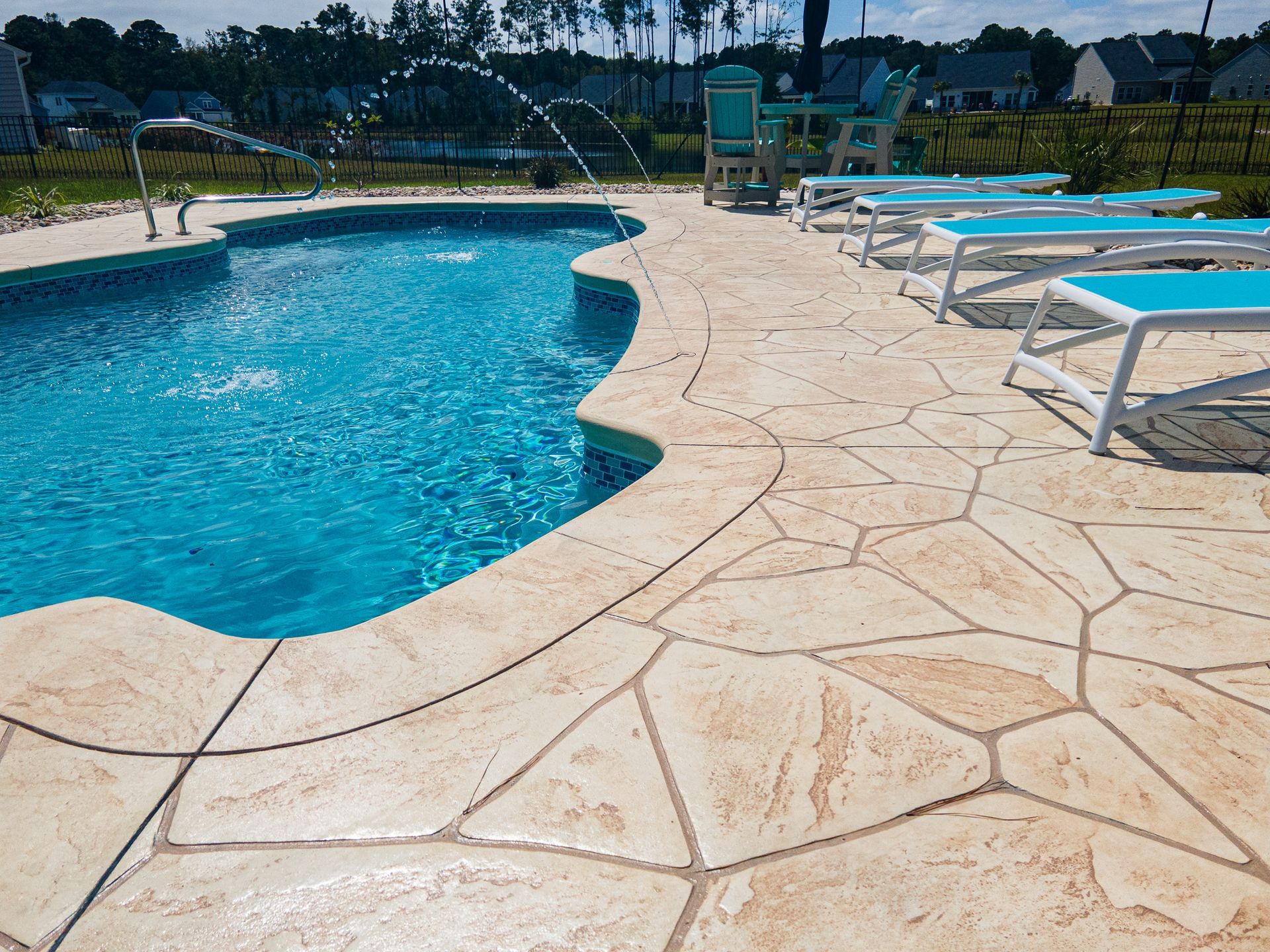 Curved blue swimming pool beside a tan stone patio with lounge chairs in a sunny outdoor setting