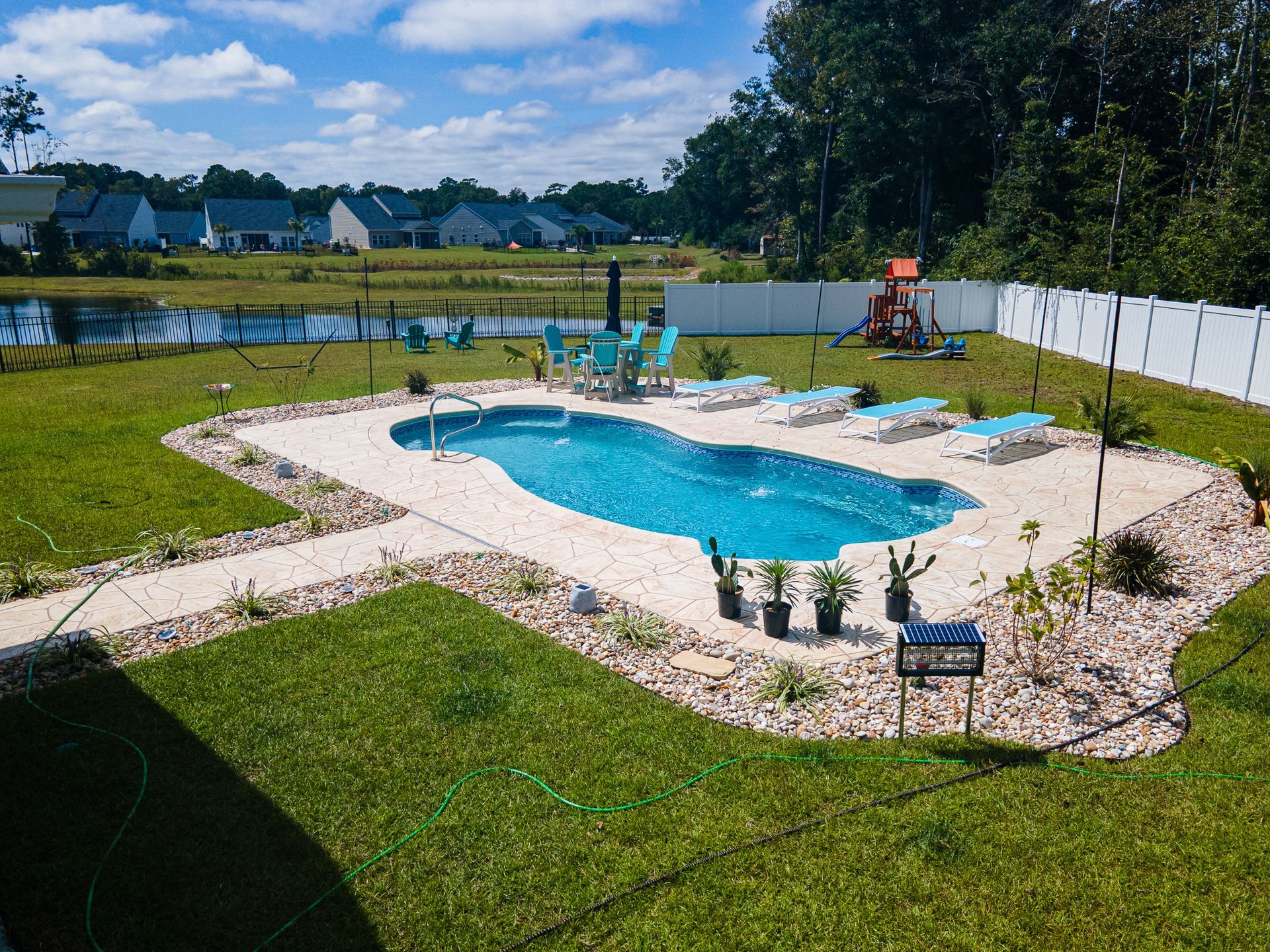 Backyard pool with stone patio, green lawn, lounge chairs, and white fence on a sunny day