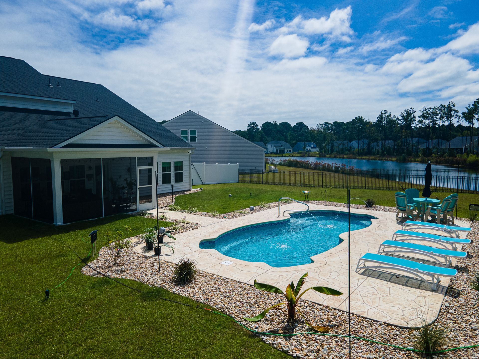 Backyard pool beside a house with blue lounge chairs, green lawn, and lake view under a cloudy sky