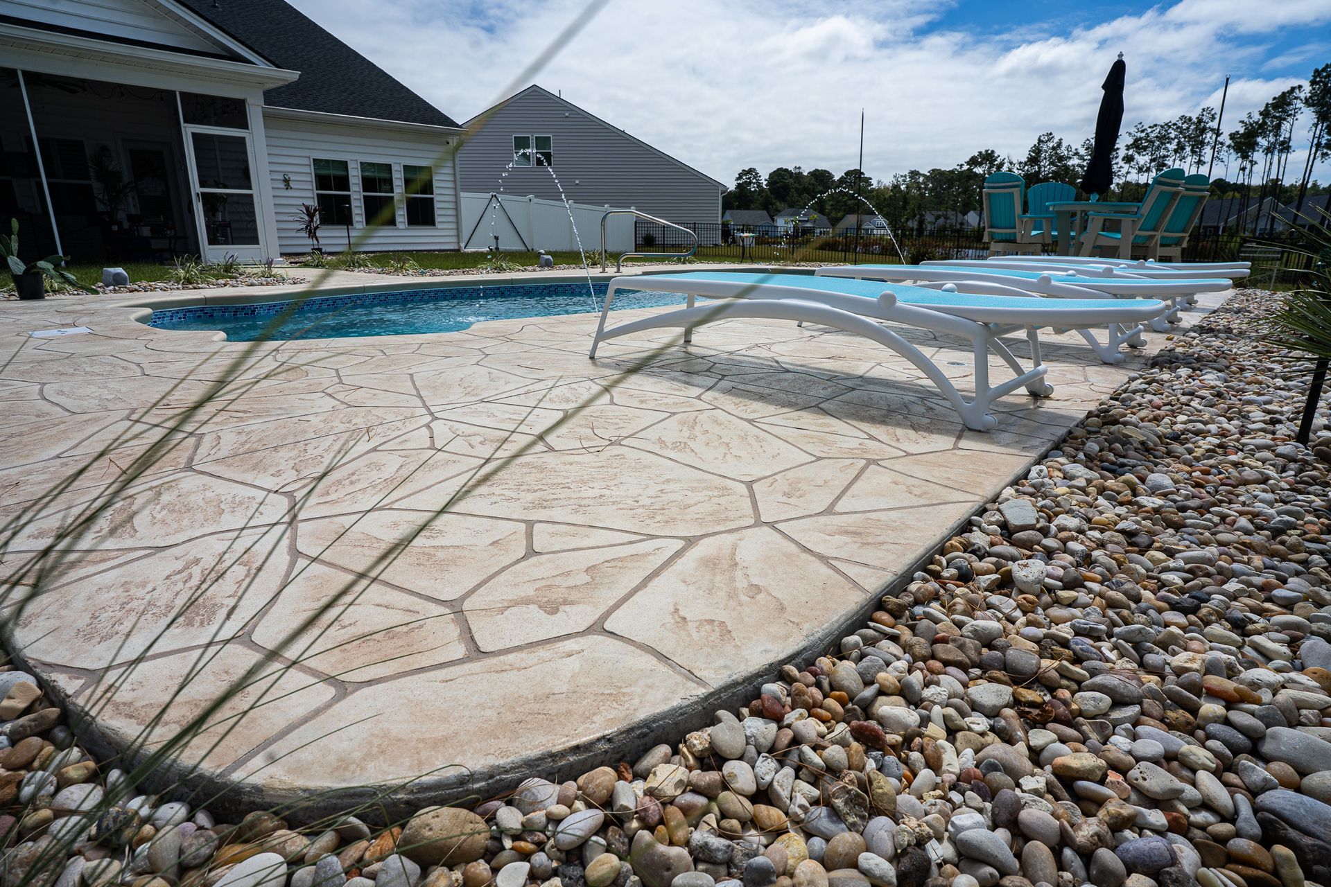Backyard pool with white lounge chairs, stone patio, and pebbled landscaping beside a house