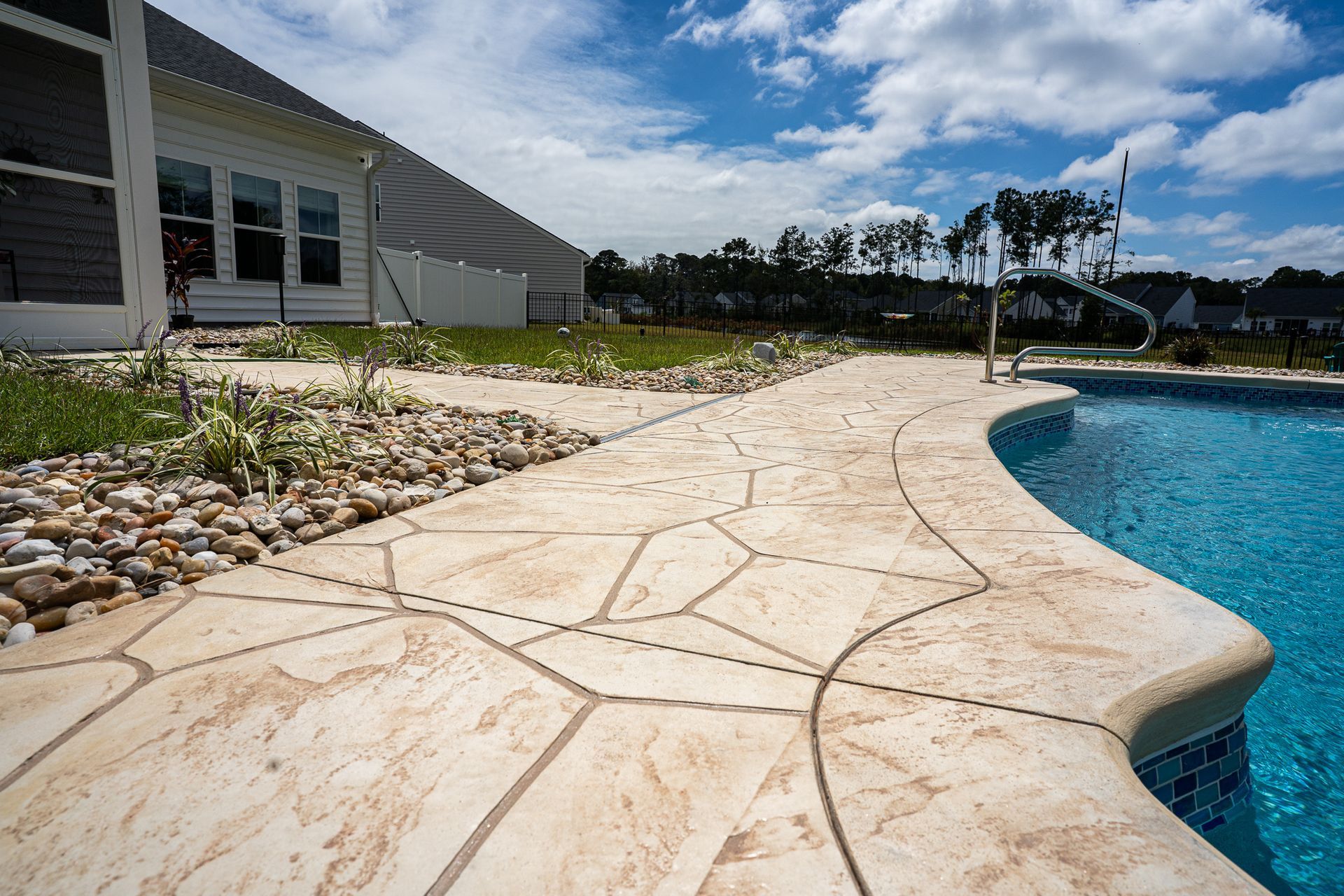 Stone pool deck beside a modern building, curving around a blue swimming pool under a cloudy sky