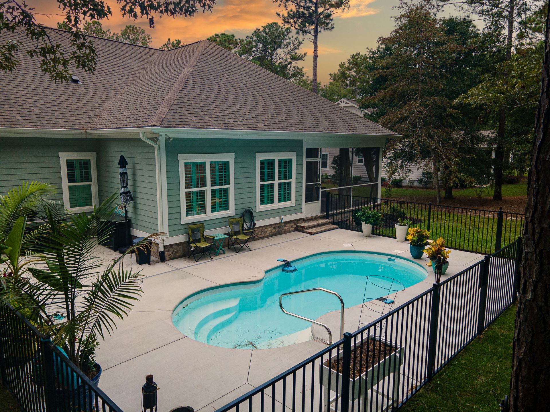 Backyard pool beside a teal house at sunset, with black fence and patio seating.