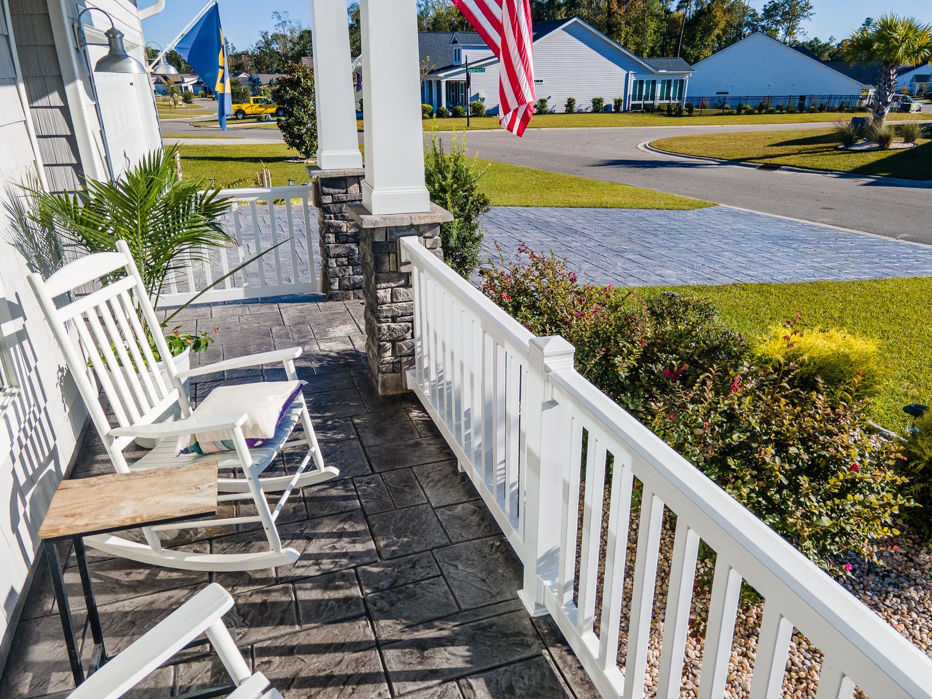 Front porch with white railings, a chair, stone steps, and an American flag overlooking a sunny neighborhood.