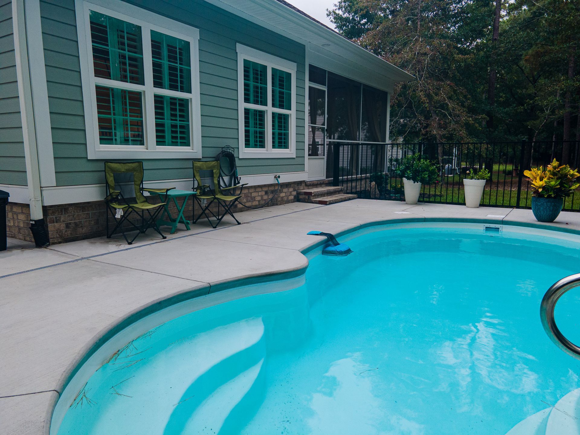 Backyard pool beside a green house with patio chairs and potted plants