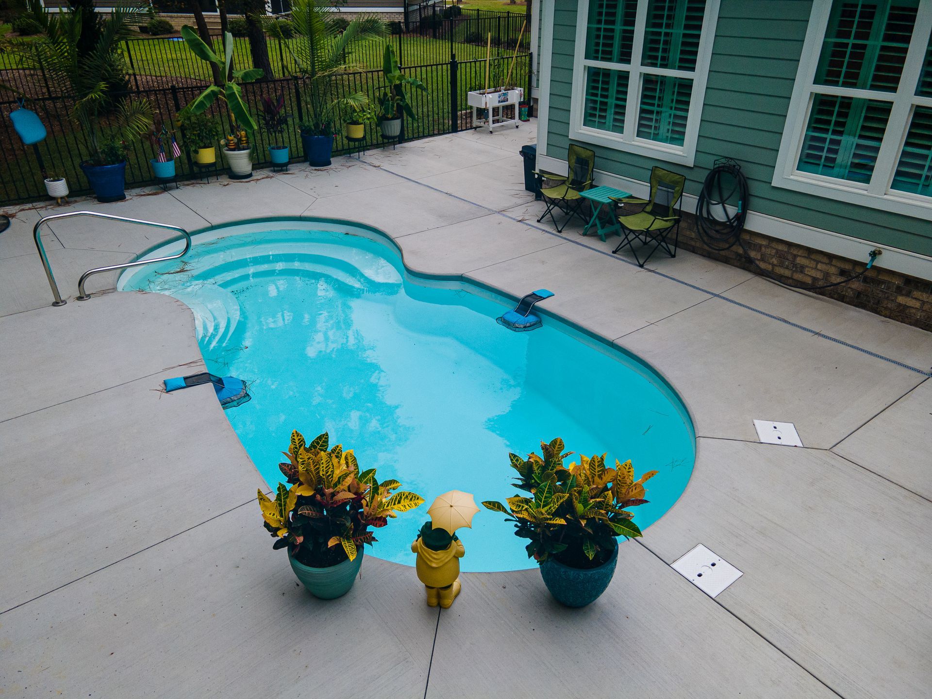 Backyard pool with turquoise water, patio pavers, and potted plants beside a house with green shutters