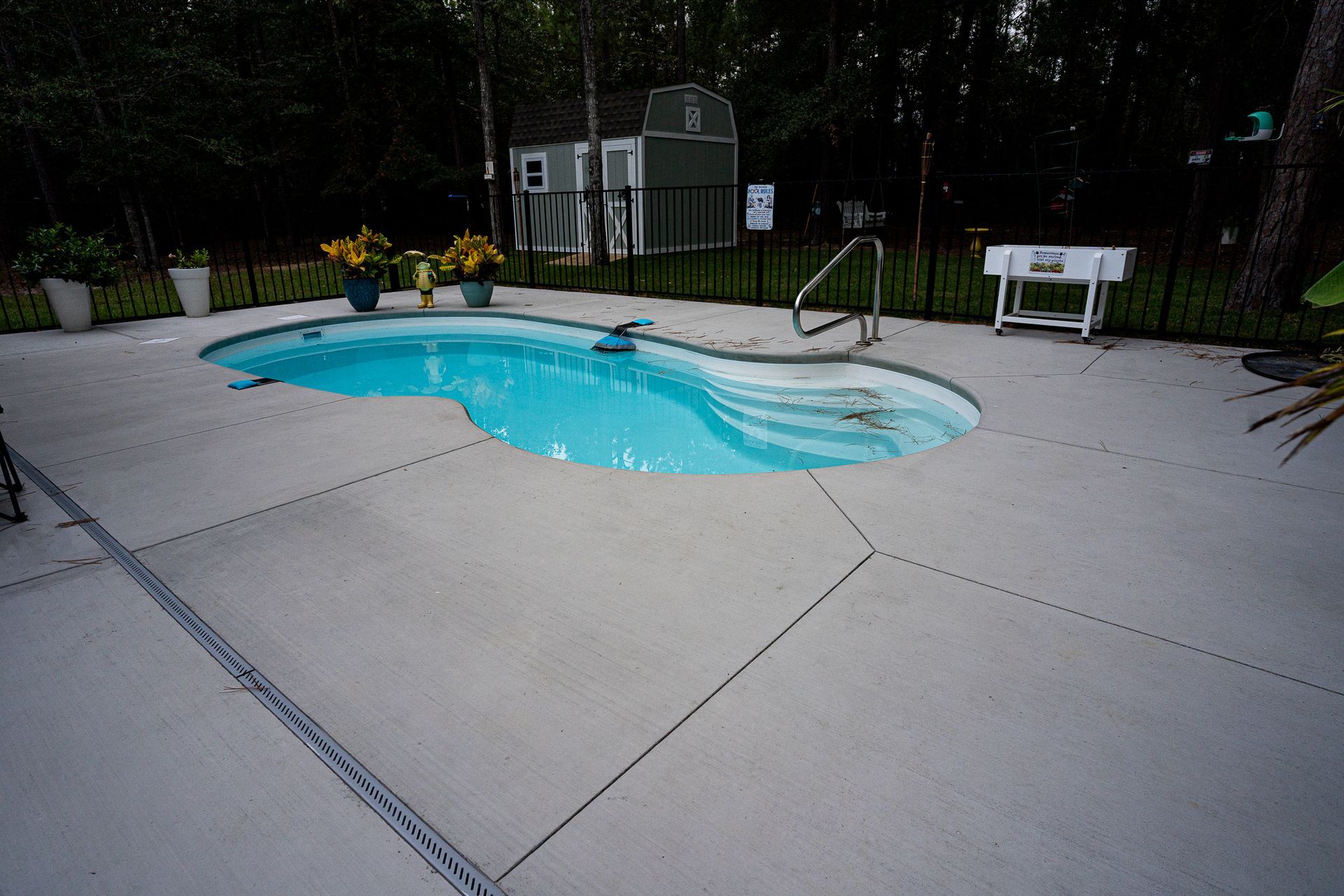 Backyard swimming pool with blue water, concrete patio, lounge chair, and greenery at dusk