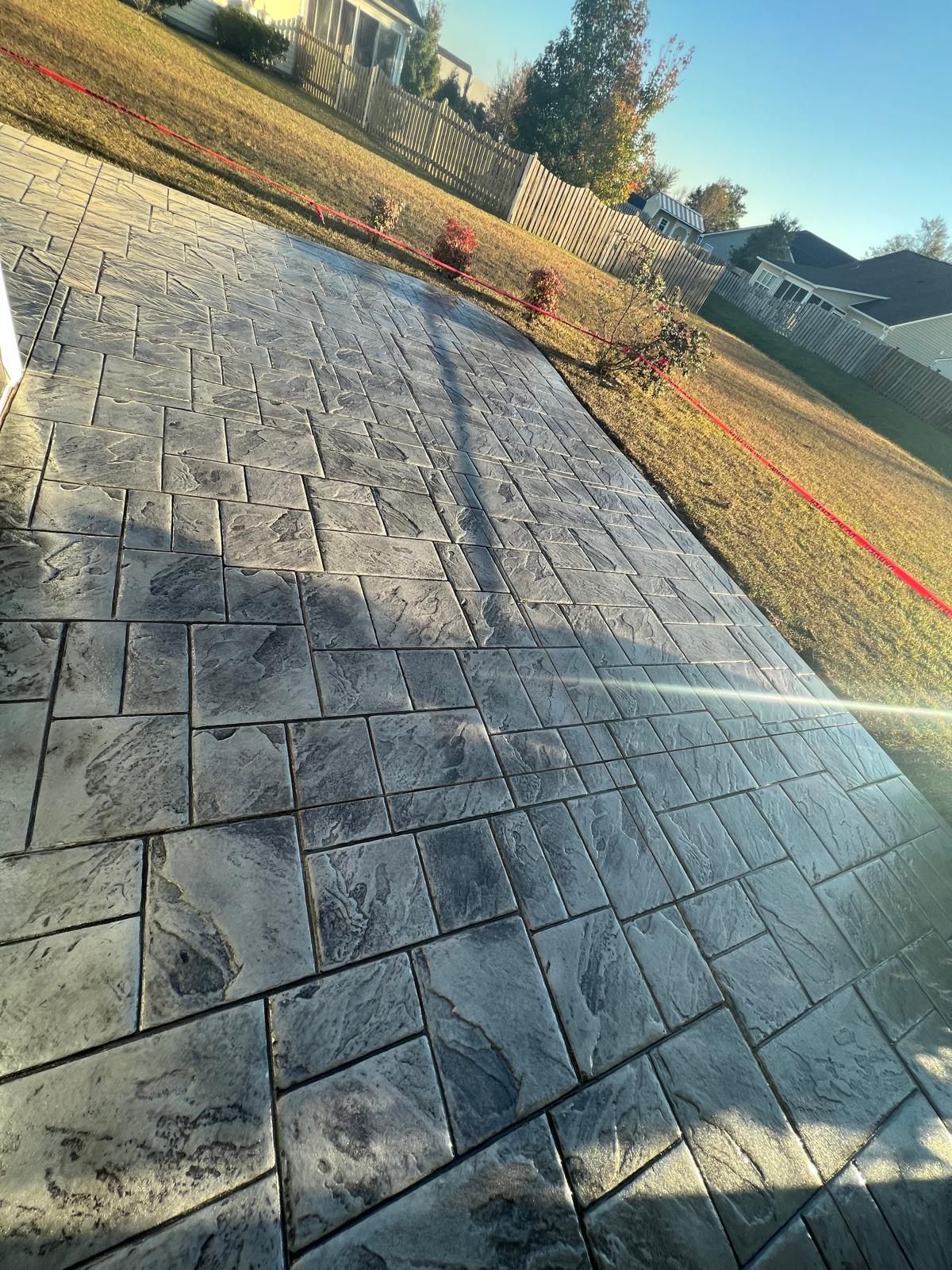Stone-paved driveway with long shadows, a red hose, and a fenced yard in warm sunlight